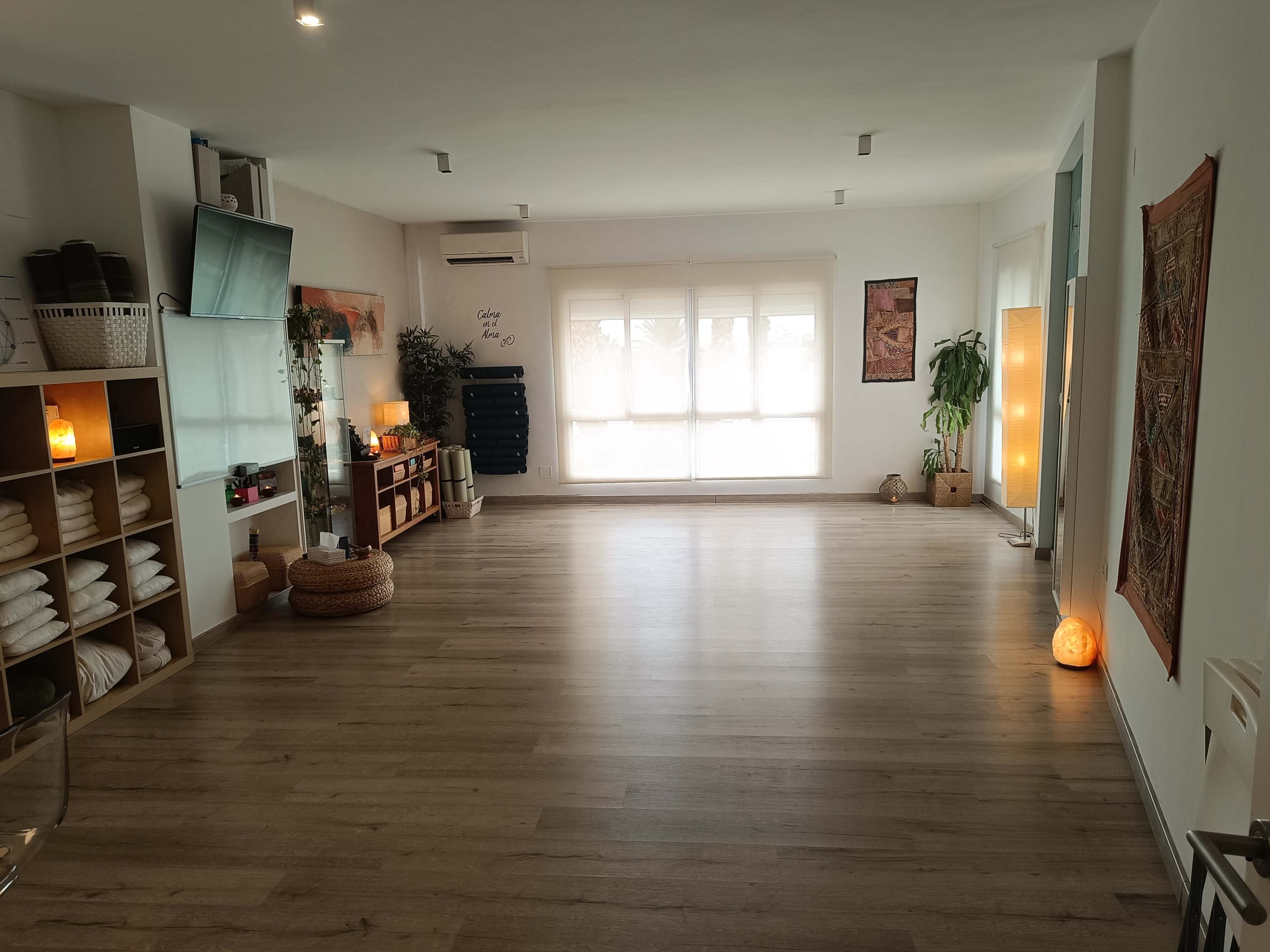 The image shows a spacious and minimally decorated room with wooden flooring, large windows, and shelves containing rolled towels and decorative elements.