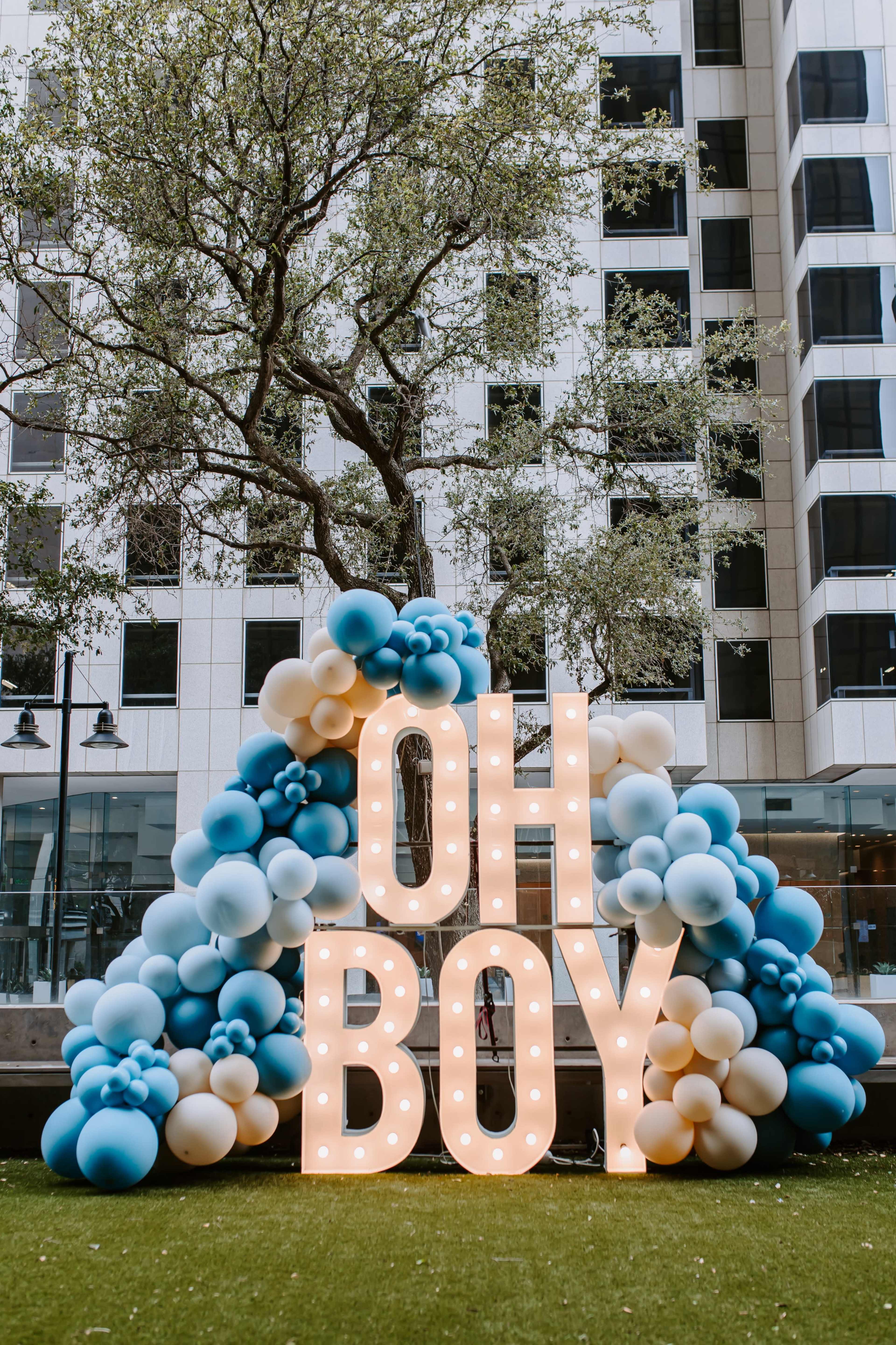 A colorful balloon arch displaying the words "OH BOY" in illuminated letters is set against a backdrop of tall buildings and trees.