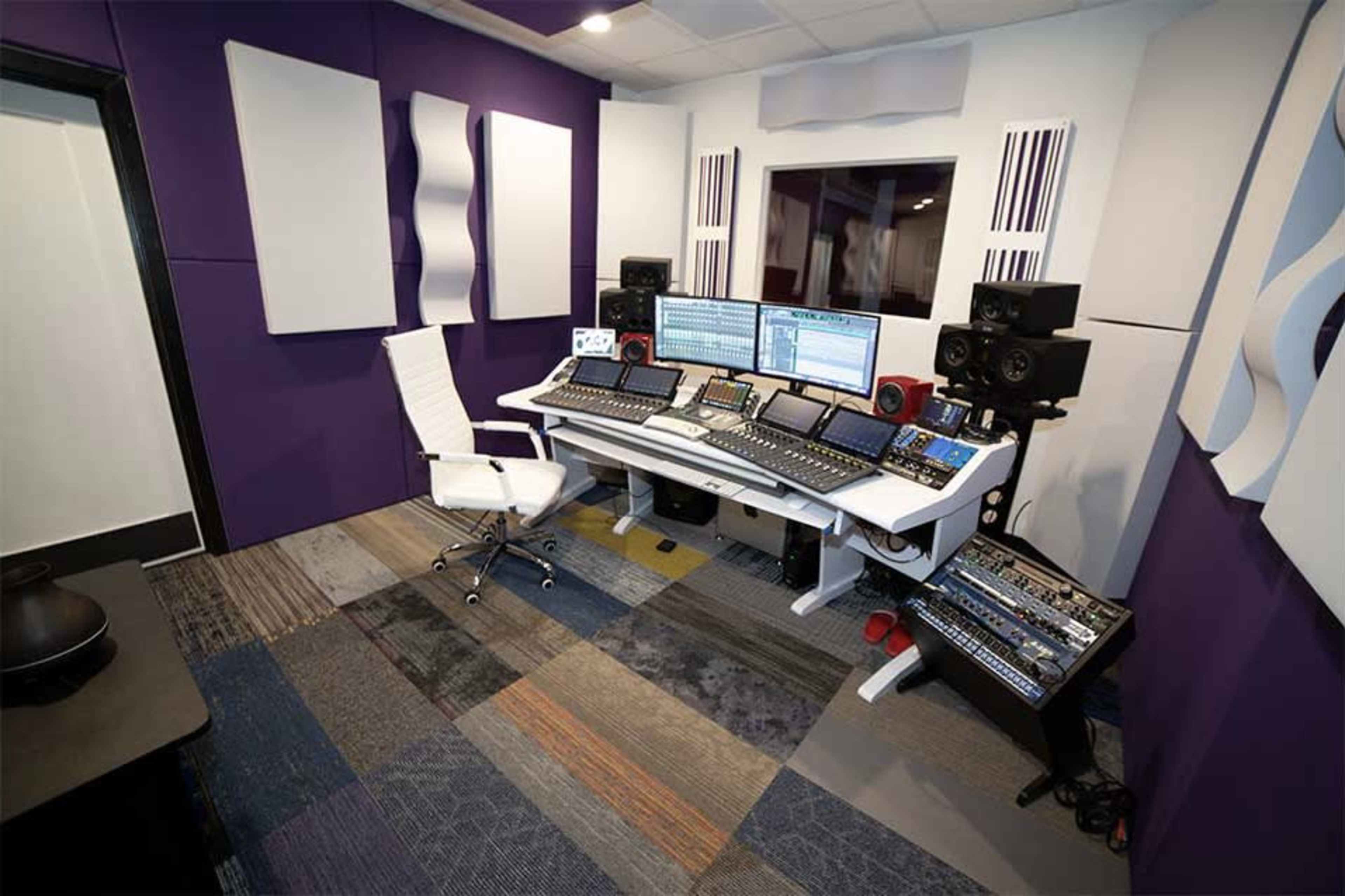 A modern music production studio with a large desk equipped with multiple digital audio workstations and audio equipment, surrounded by purple acoustic panels.