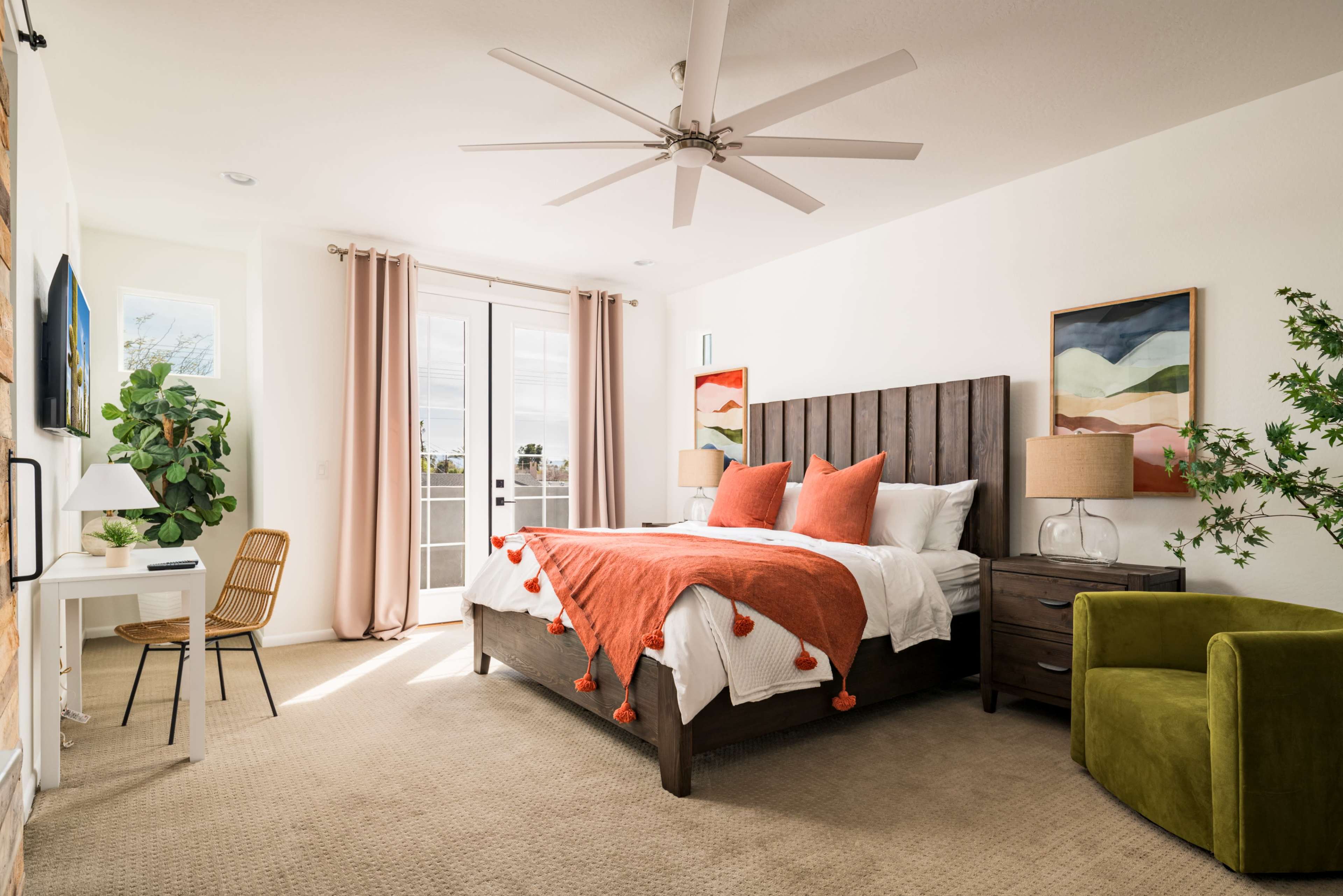 The image shows a bright bedroom featuring a large bed with an orange throw blanket, a chair, a desk, and a fan, with double doors leading to a balcony.