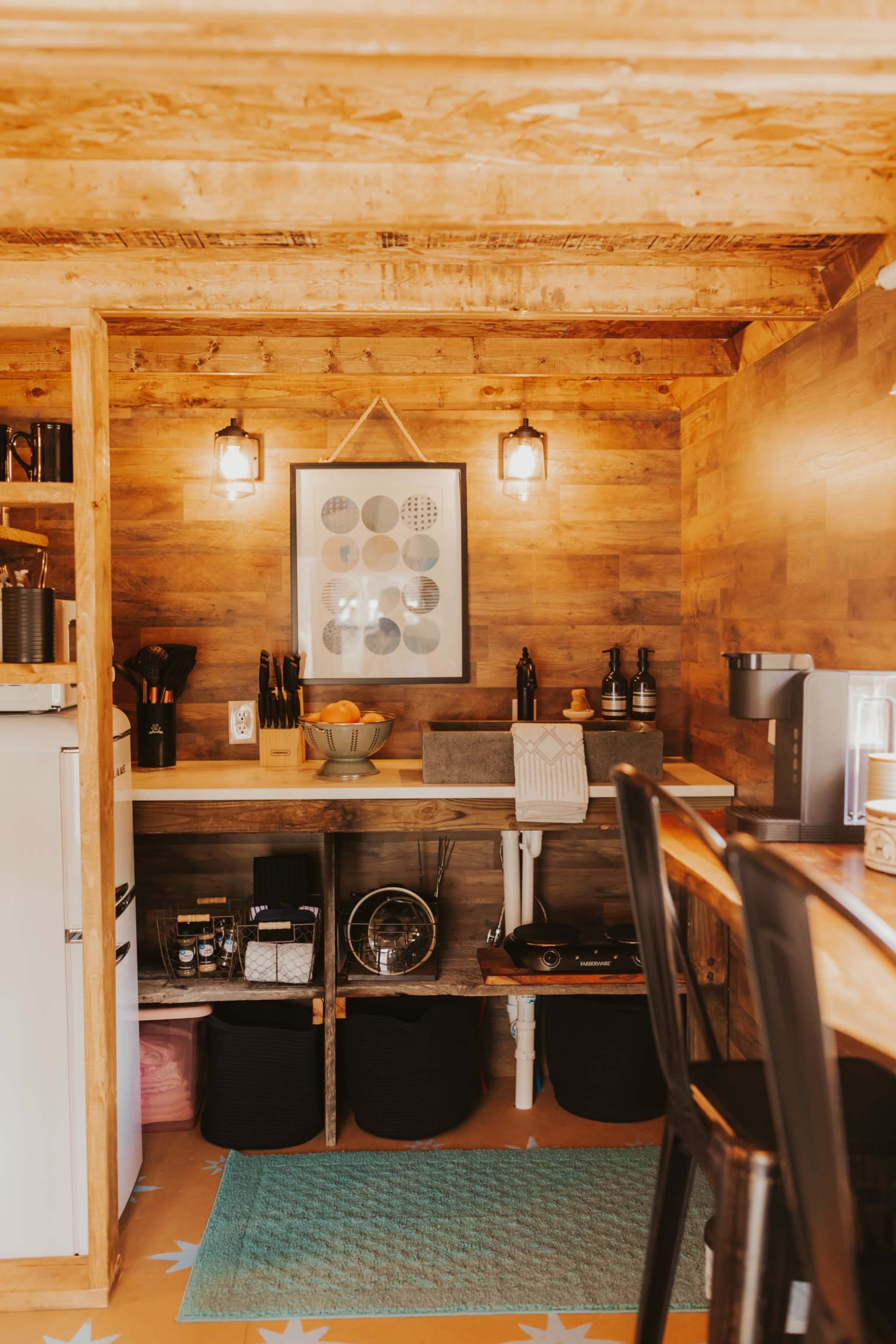 The image shows a small kitchen area with wooden walls and a countertop featuring various kitchen items, including a fruit bowl and kitchen appliances.