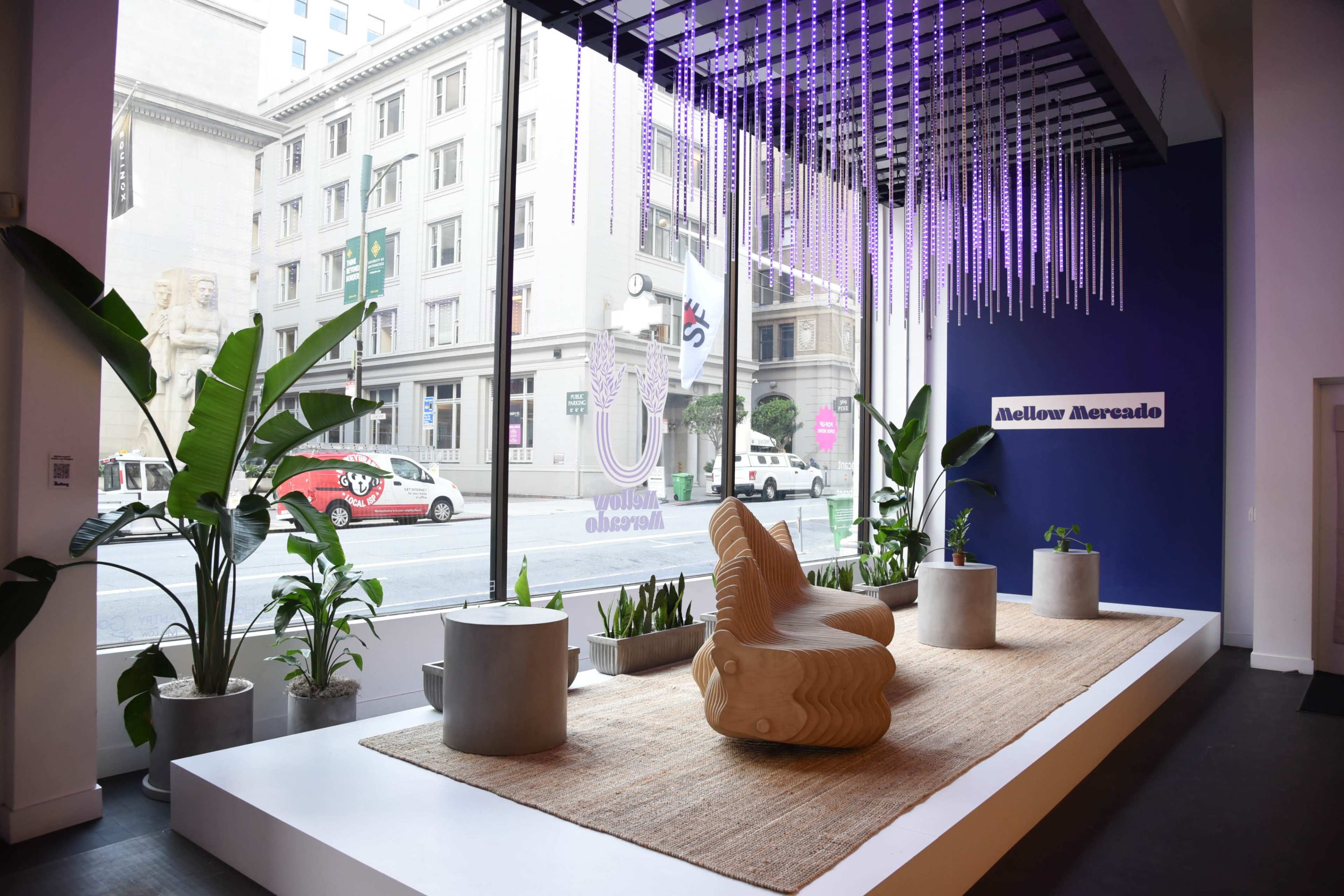 A modern interior space with a wooden chair, potted plants, and decorative lighting, situated by large windows overlooking a city street.