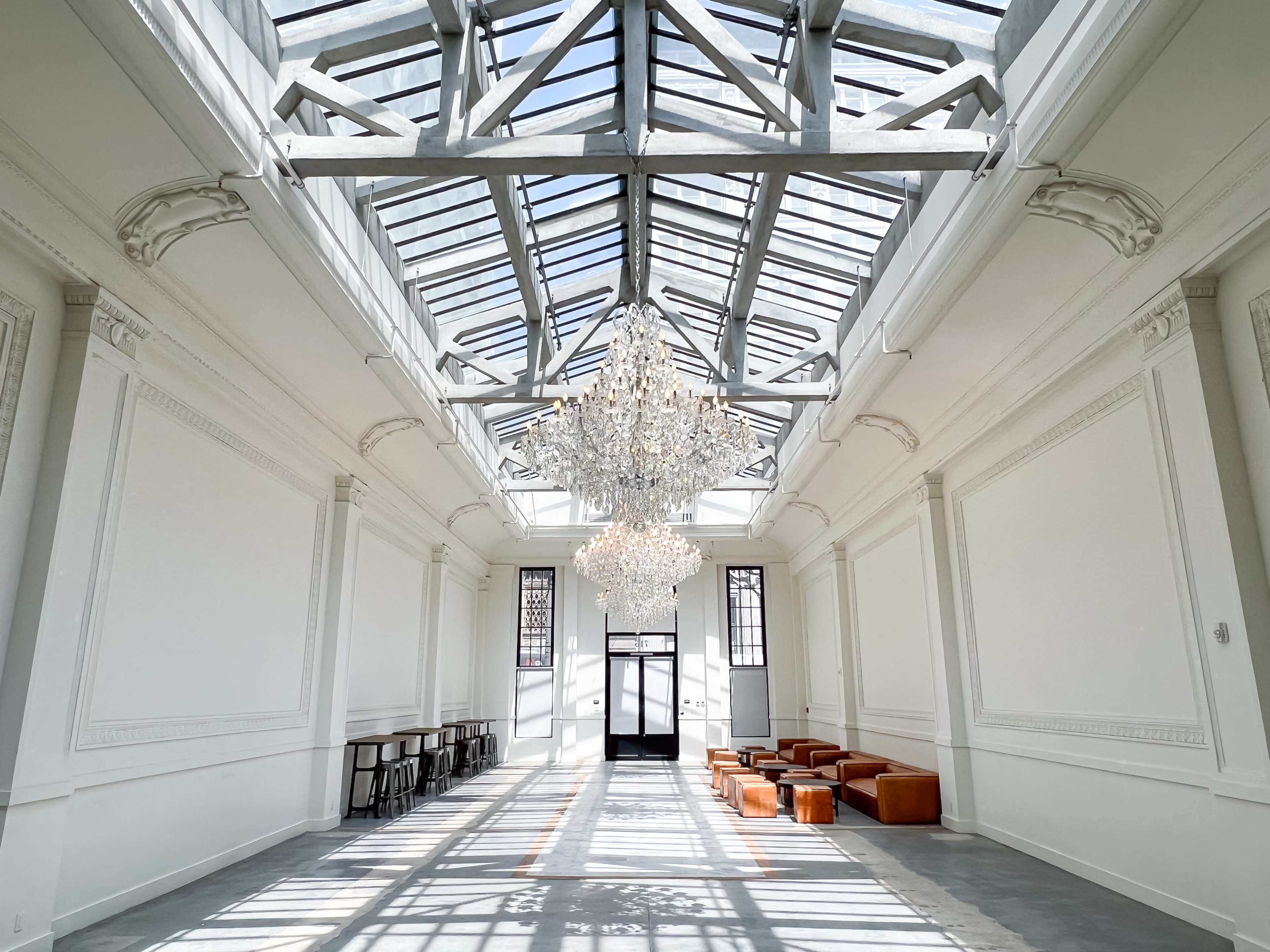 A spacious interior with a high glass roof, featuring a chandelier, along with leather seating and a set of metal chairs along one side.