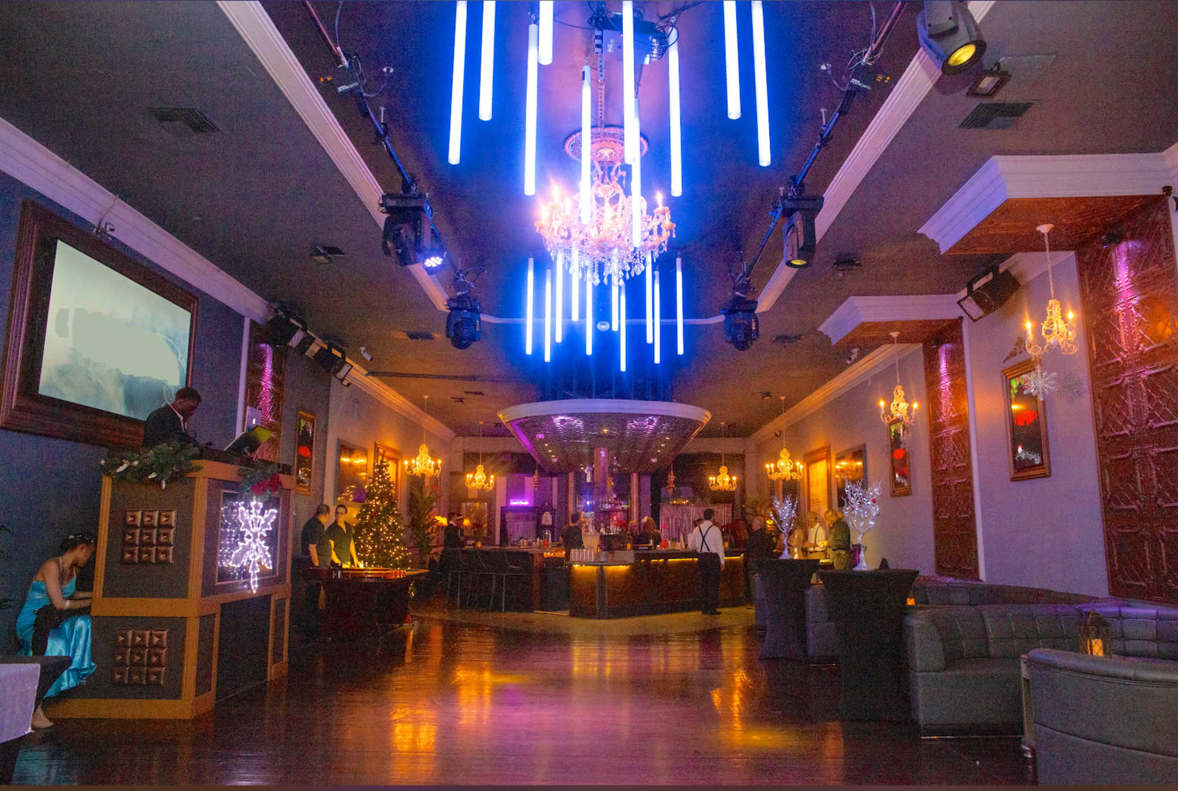 A modern lounge interior features a central bar with glowing neon lights and decorative chandeliers, surrounded by elegant seating and artwork on the walls.