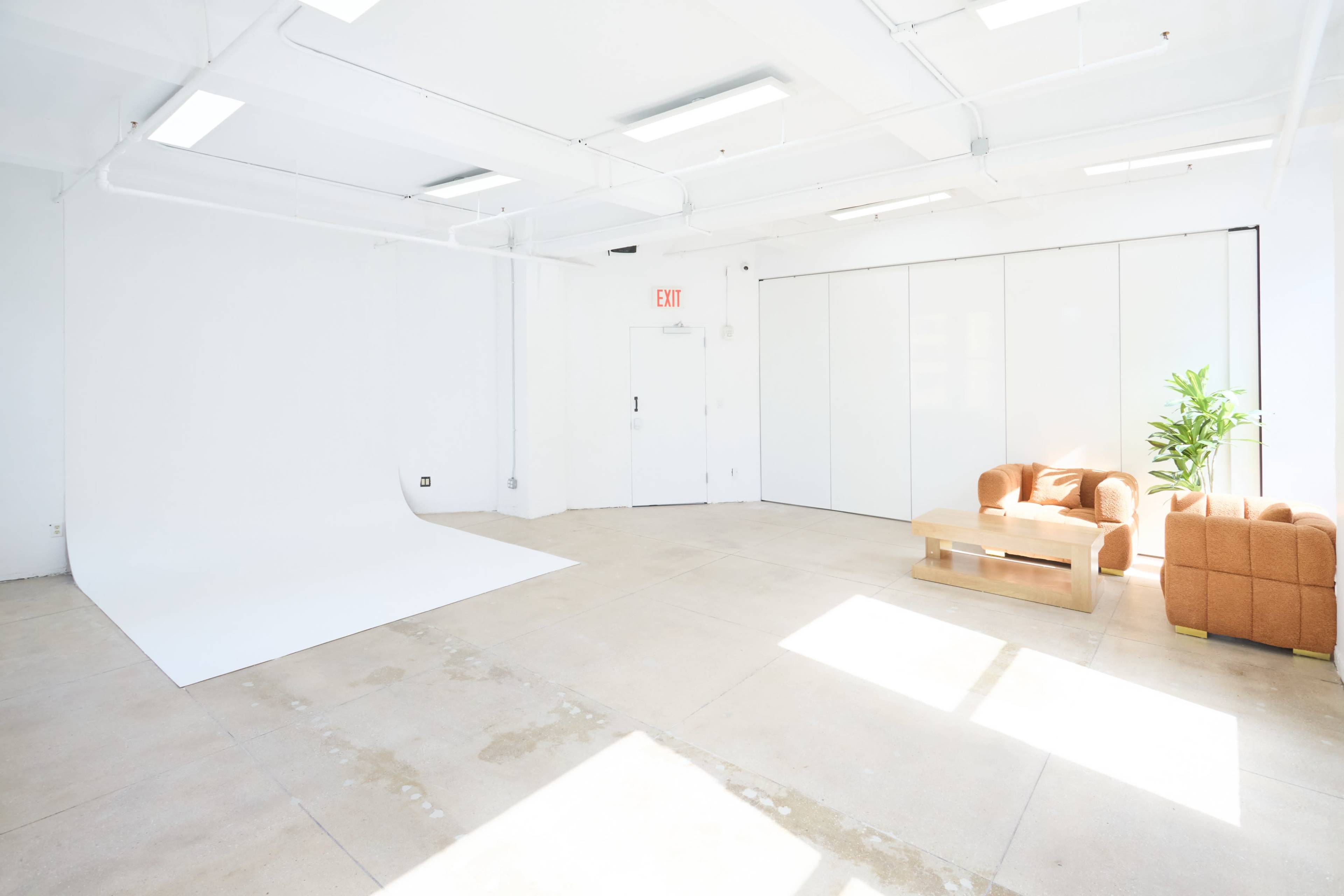 The image shows a bright, spacious studio with a white backdrop, a wooden coffee table, and a brown couch positioned near a large window.