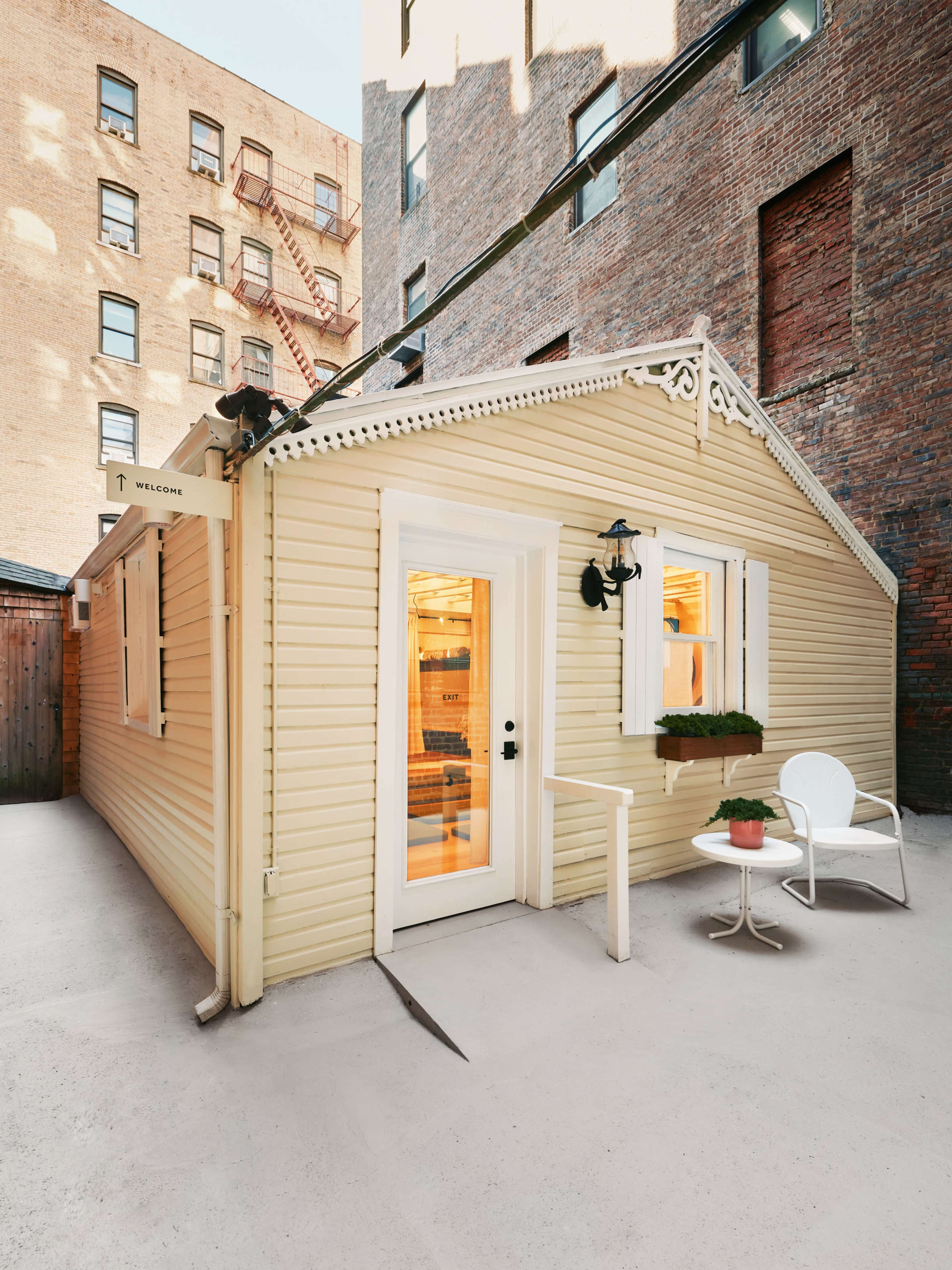 The image shows a small, yellow cottage with a white door and a chair set outside on a paved surface, surrounded by brick buildings.