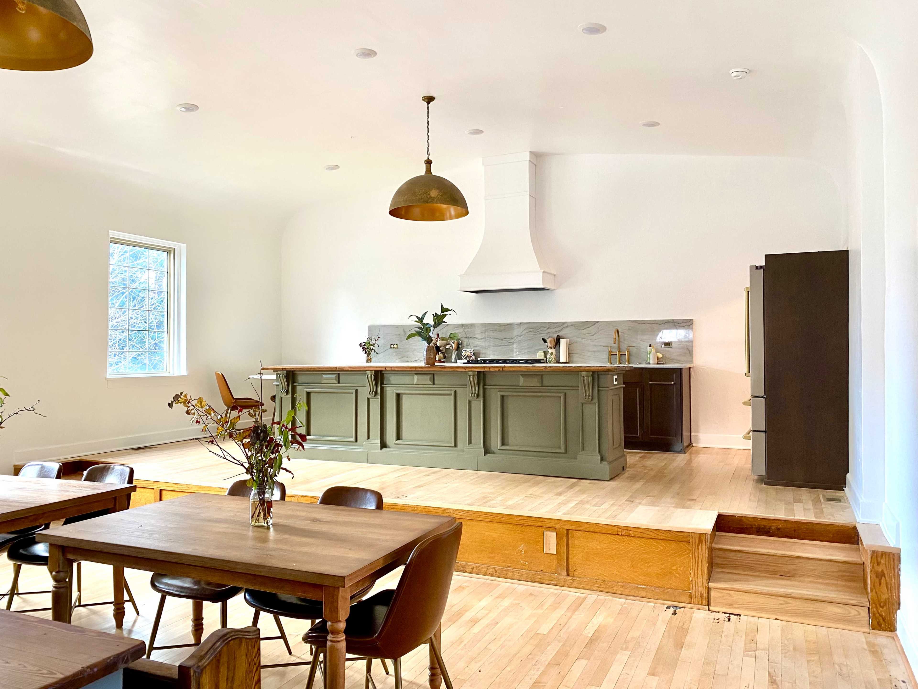 A spacious kitchen features a green island with bar seating, a large hood above the stove, and wooden tables set for dining.