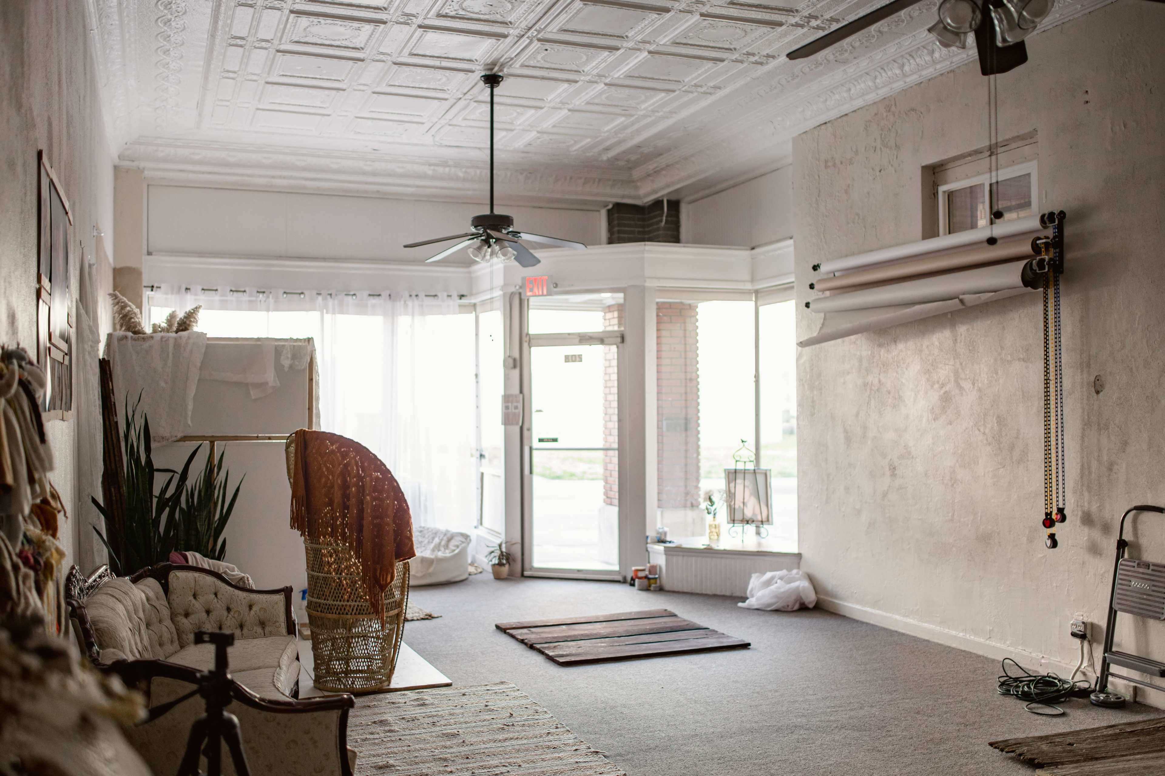 The image shows a spacious and empty room with a textured ceiling, featuring a seating area, plants, and large windows letting in natural light.