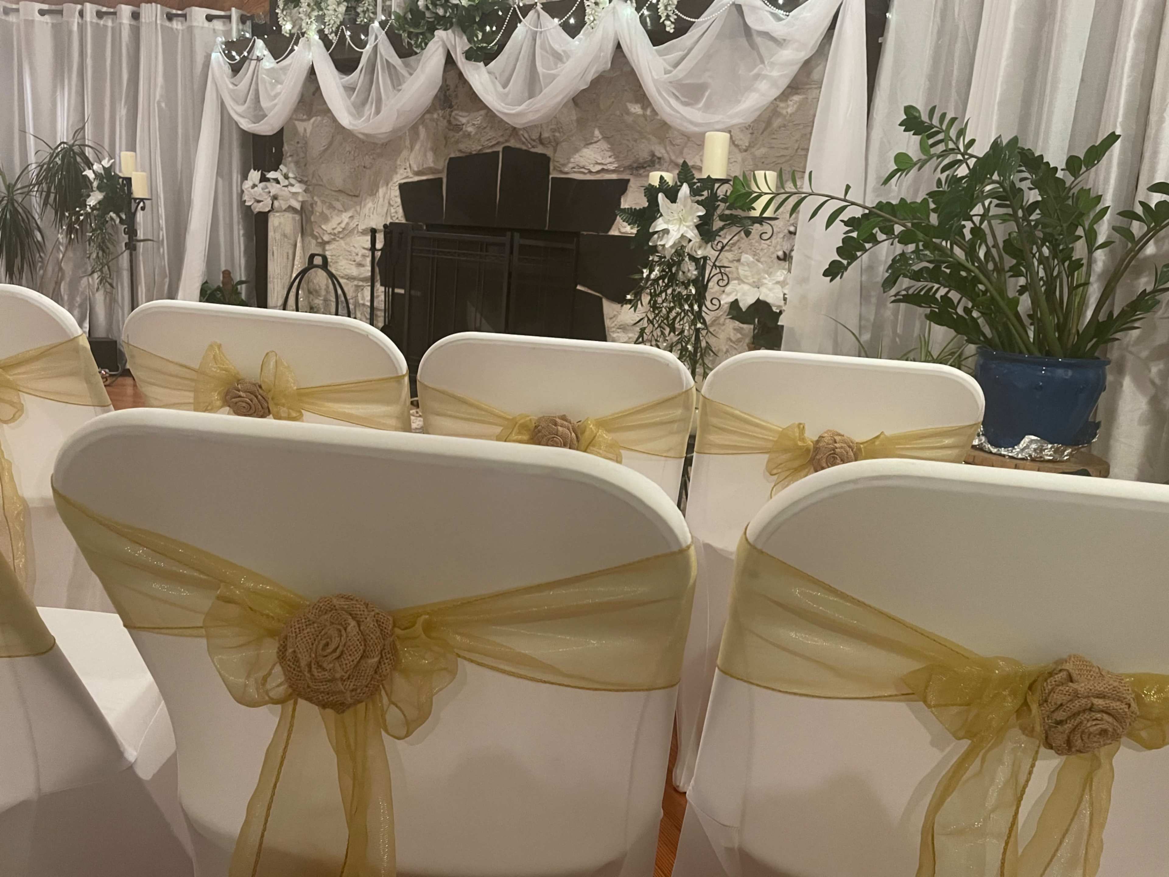 The image depicts a decorated indoor setting for a ceremony, featuring white chairs with yellow sashes arranged in rows facing a stone fireplace adorned with draped white fabric and greenery.