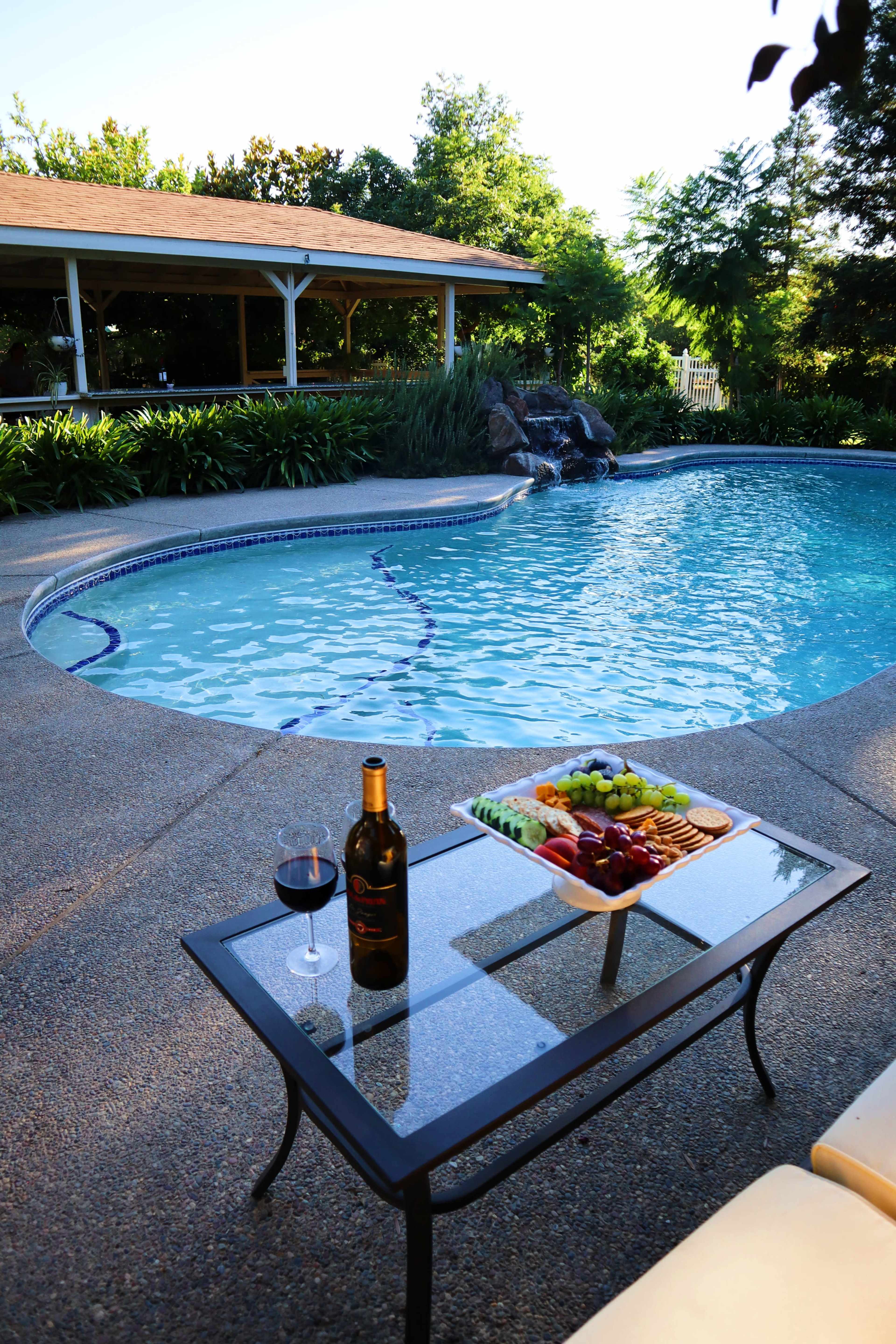 A table with a wine bottle and a platter of fruit and crackers is set beside a swimming pool with a waterfall feature.