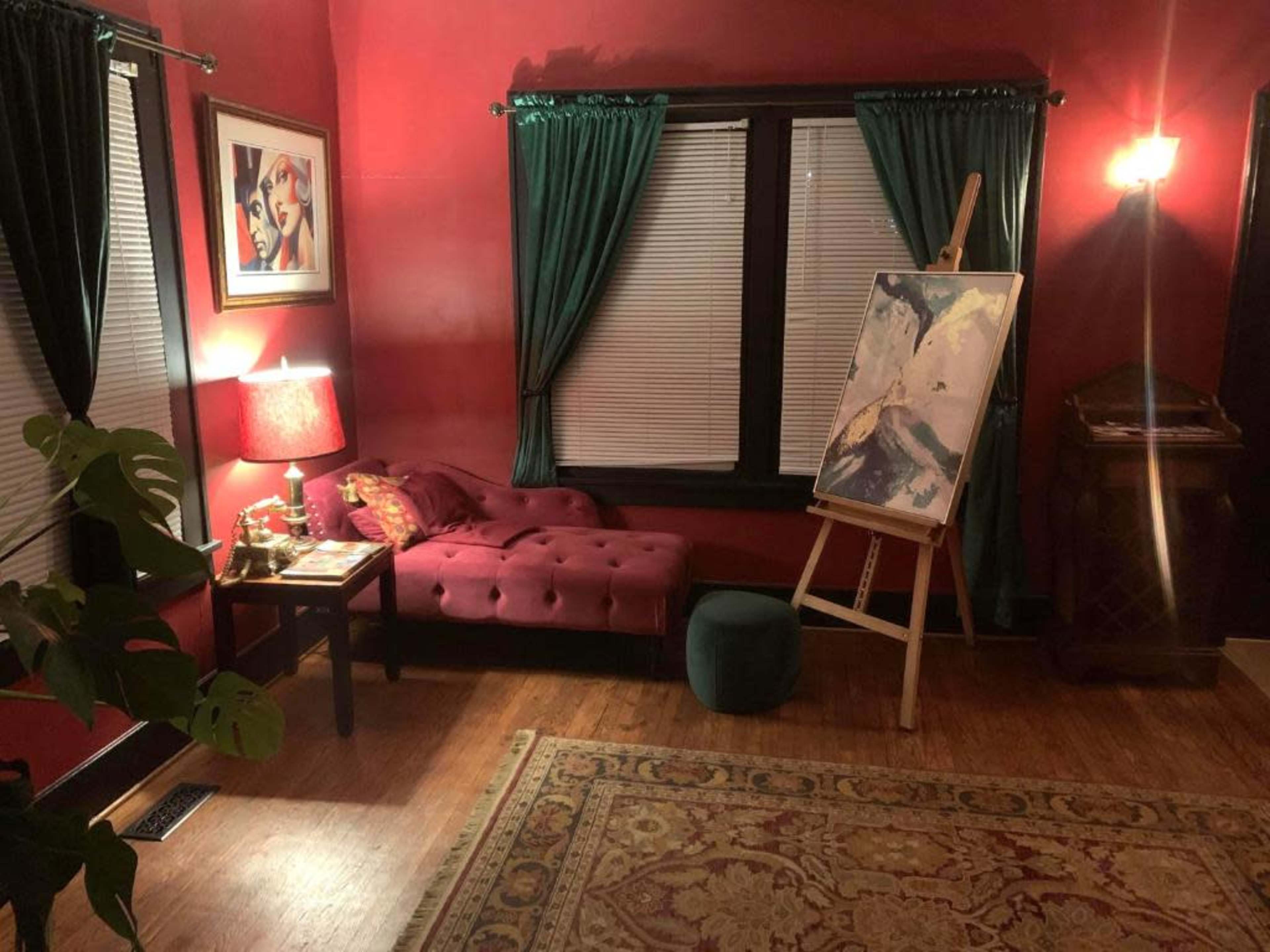 The scene features a cozy room with dark red walls, showcasing a tufted sofa, an easel with artwork, and green drapes on the windows.