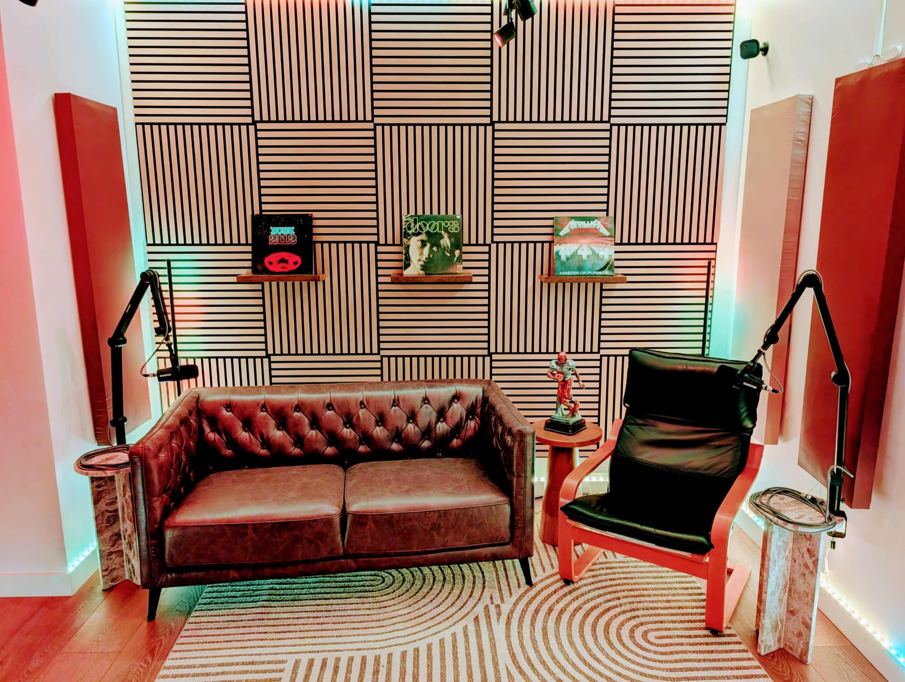 The image shows a modern audio recording studio with a tufted brown couch, a black chair, acoustic panels on the walls, and colorful lights creating a dynamic atmosphere.