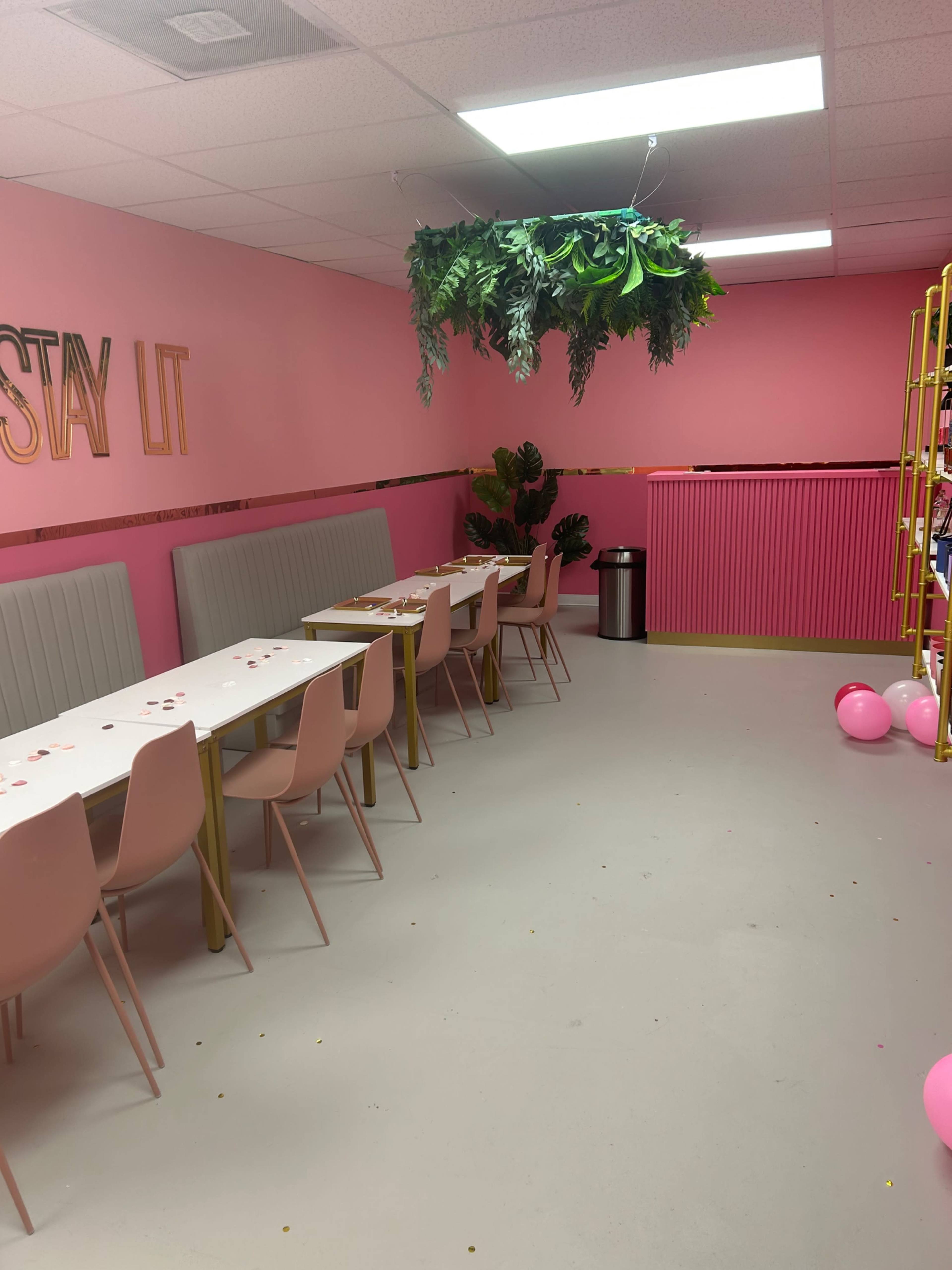 The image shows a pink-themed room with a row of pale pink chairs and tables, along with hanging greenery and decorative elements.