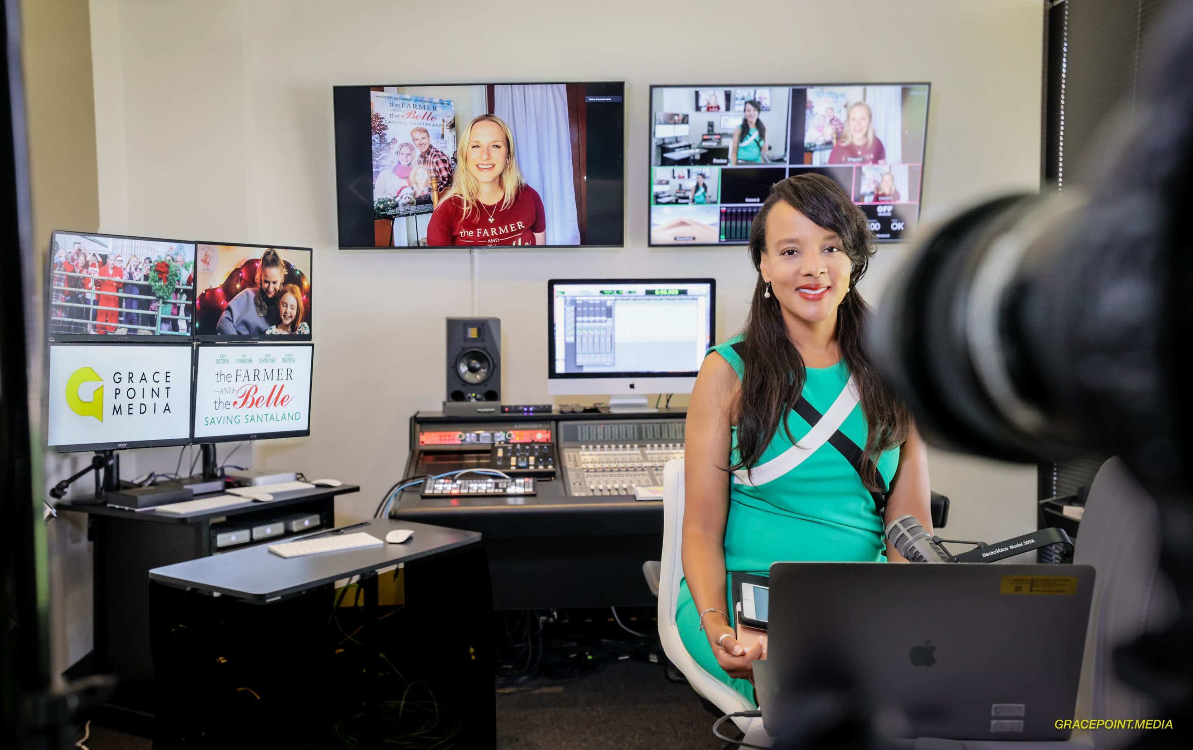 A woman in a green dress sits at a desk in a media studio, surrounded by multiple screens displaying various programs and graphics.