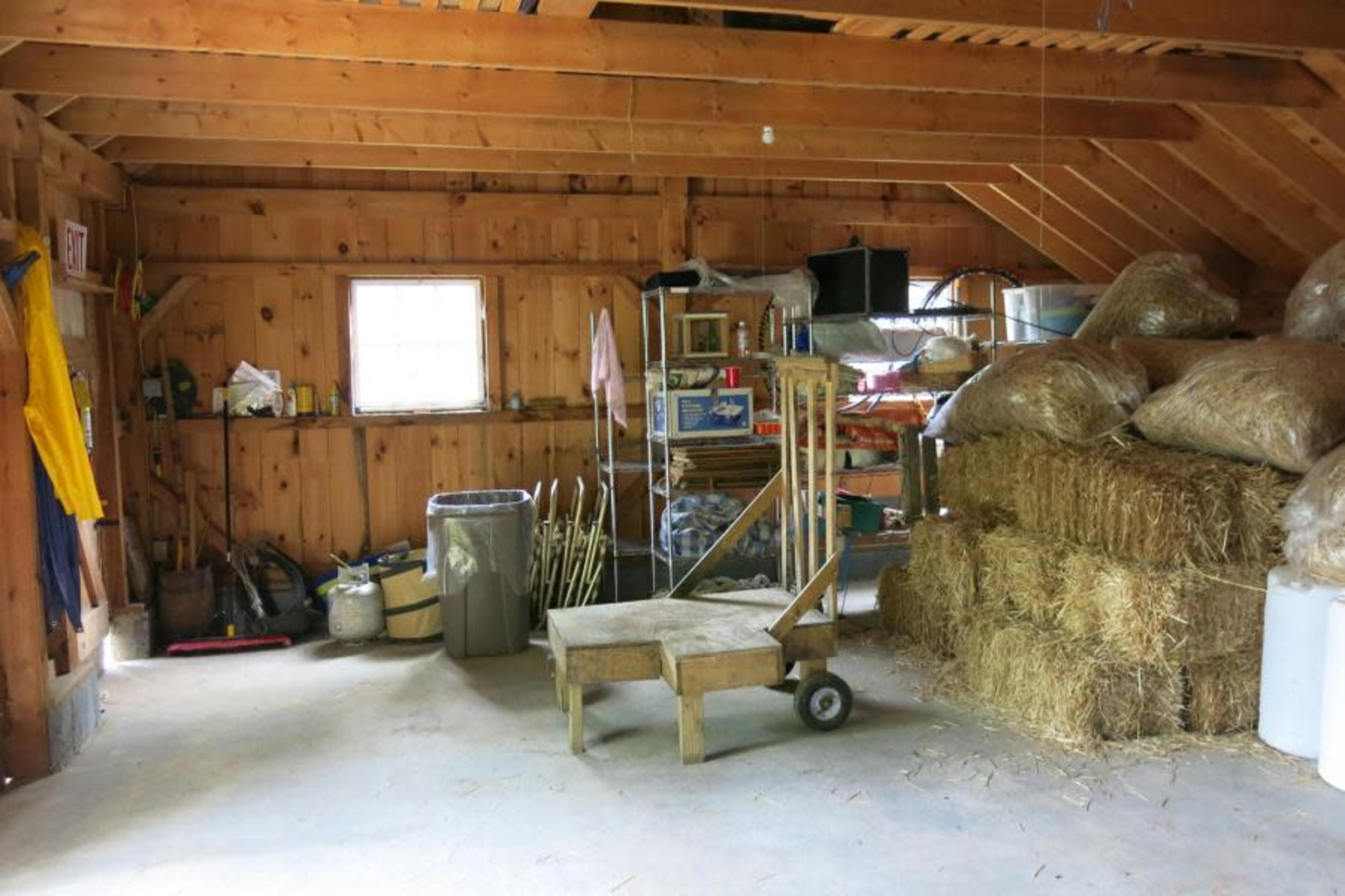The interior of a wooden barn features hay bales stacked in one corner, shelves filled with tools, and a cart in the middle of the floor.