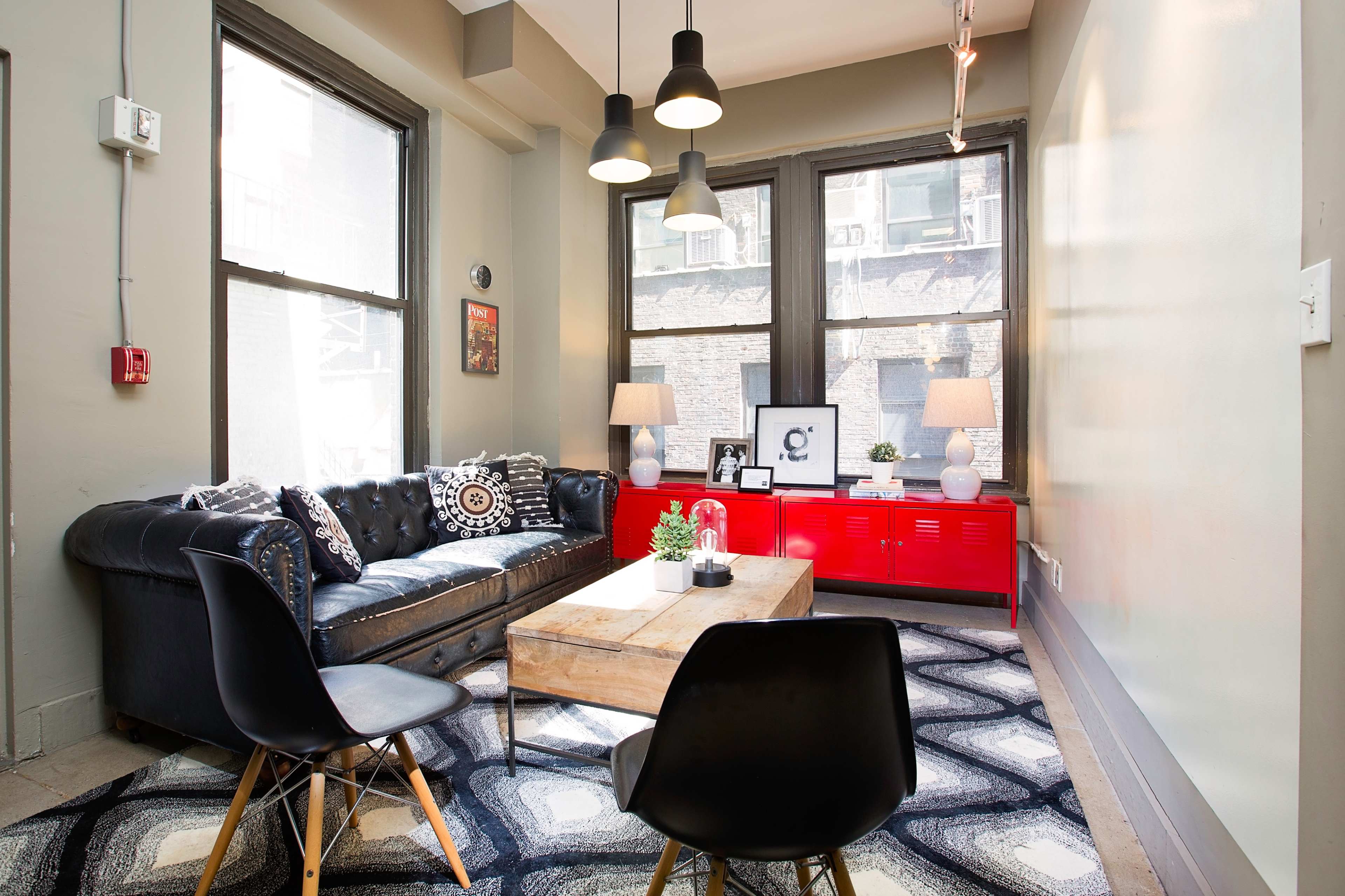 The image shows a modern living room with a black leather sofa, a wooden coffee table, and two chairs, featuring large windows that let in natural light.