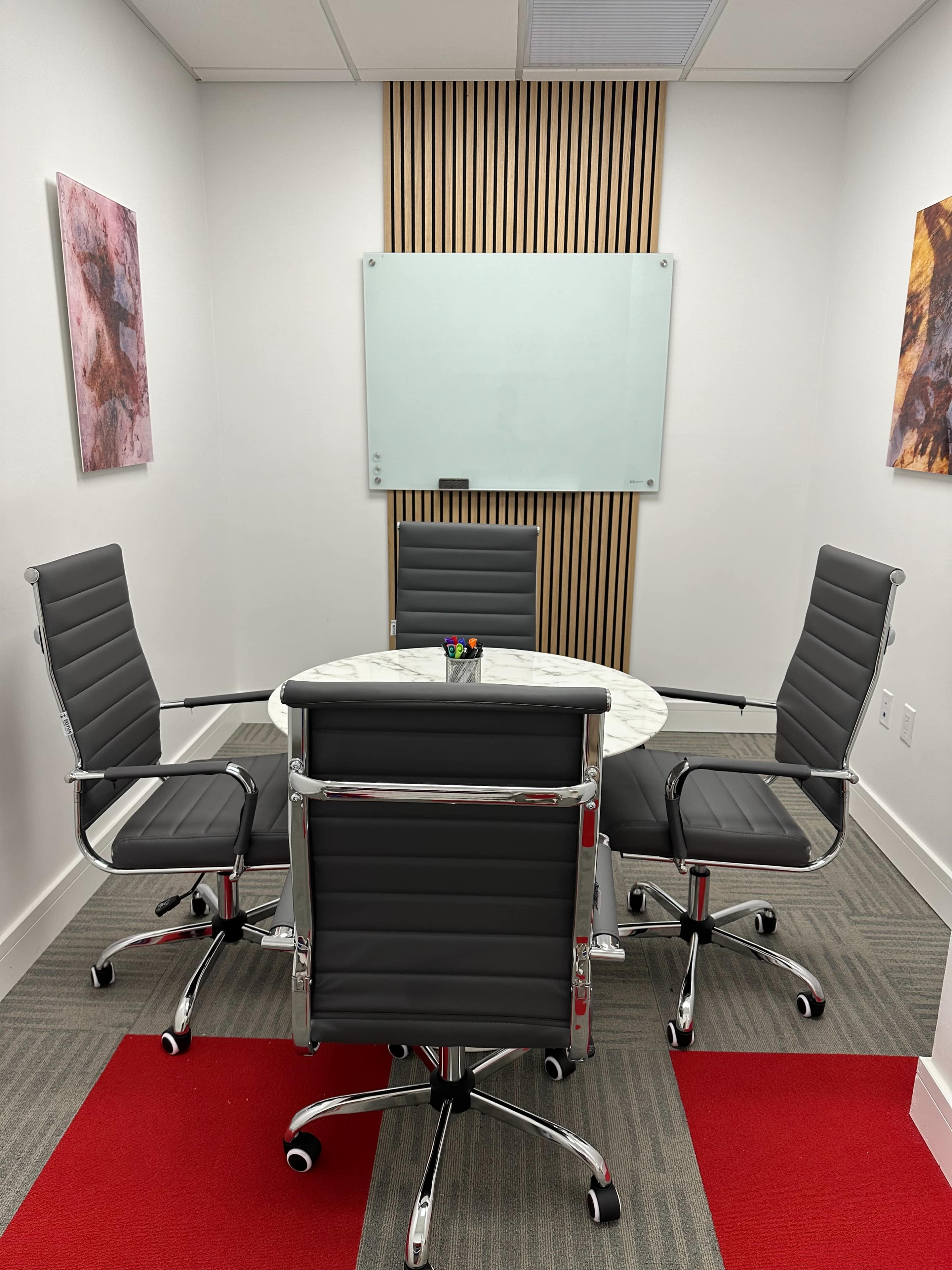 A small conference room with a round marble table and four black leather chairs, flanked by white walls and wooden paneling.