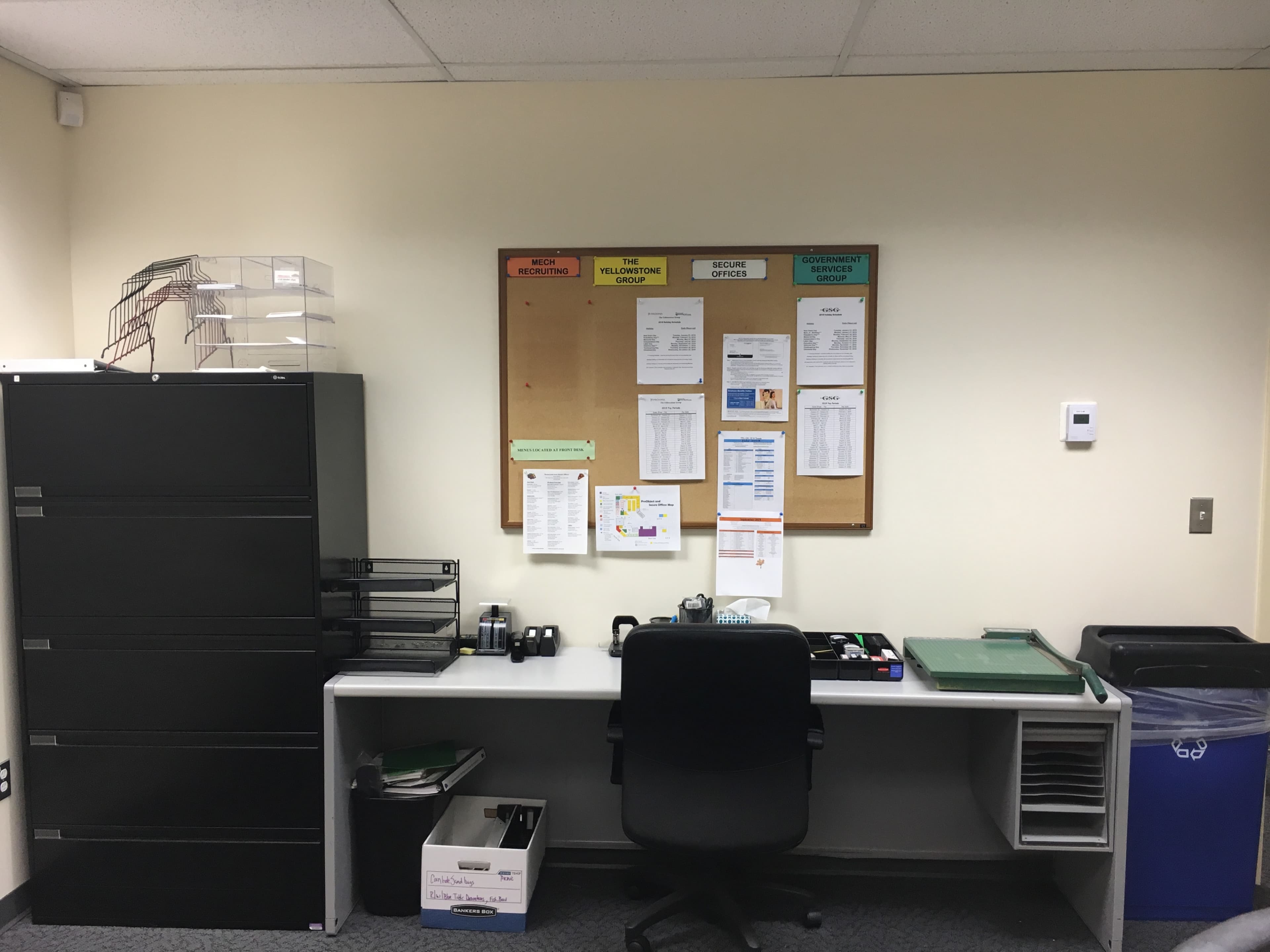 The image shows a small office with a desk, an office chair, a filing cabinet, and a bulletin board displaying various documents and notices.