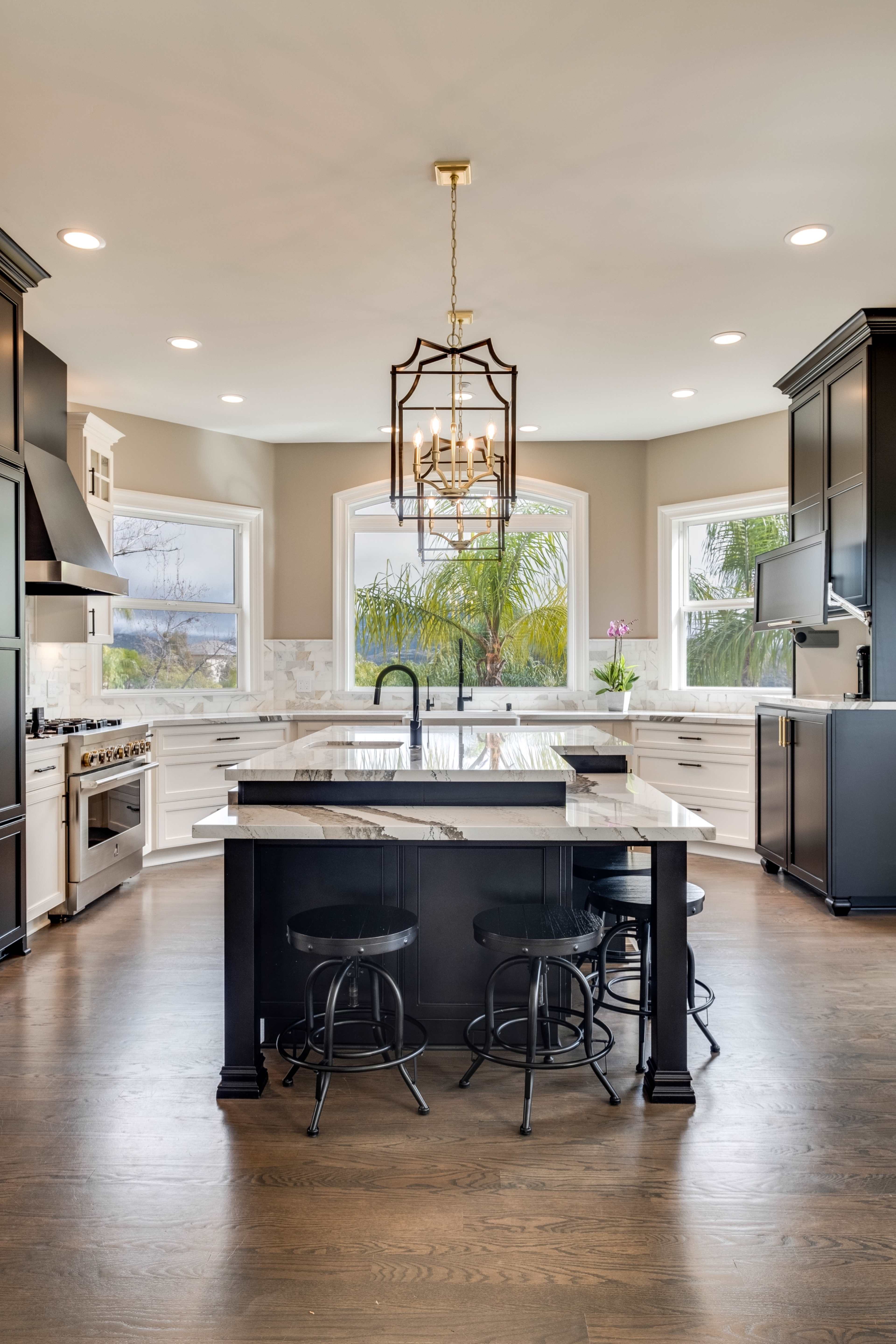 A modern kitchen features a central island with bar stools, white cabinetry, and large windows overlooking greenery.