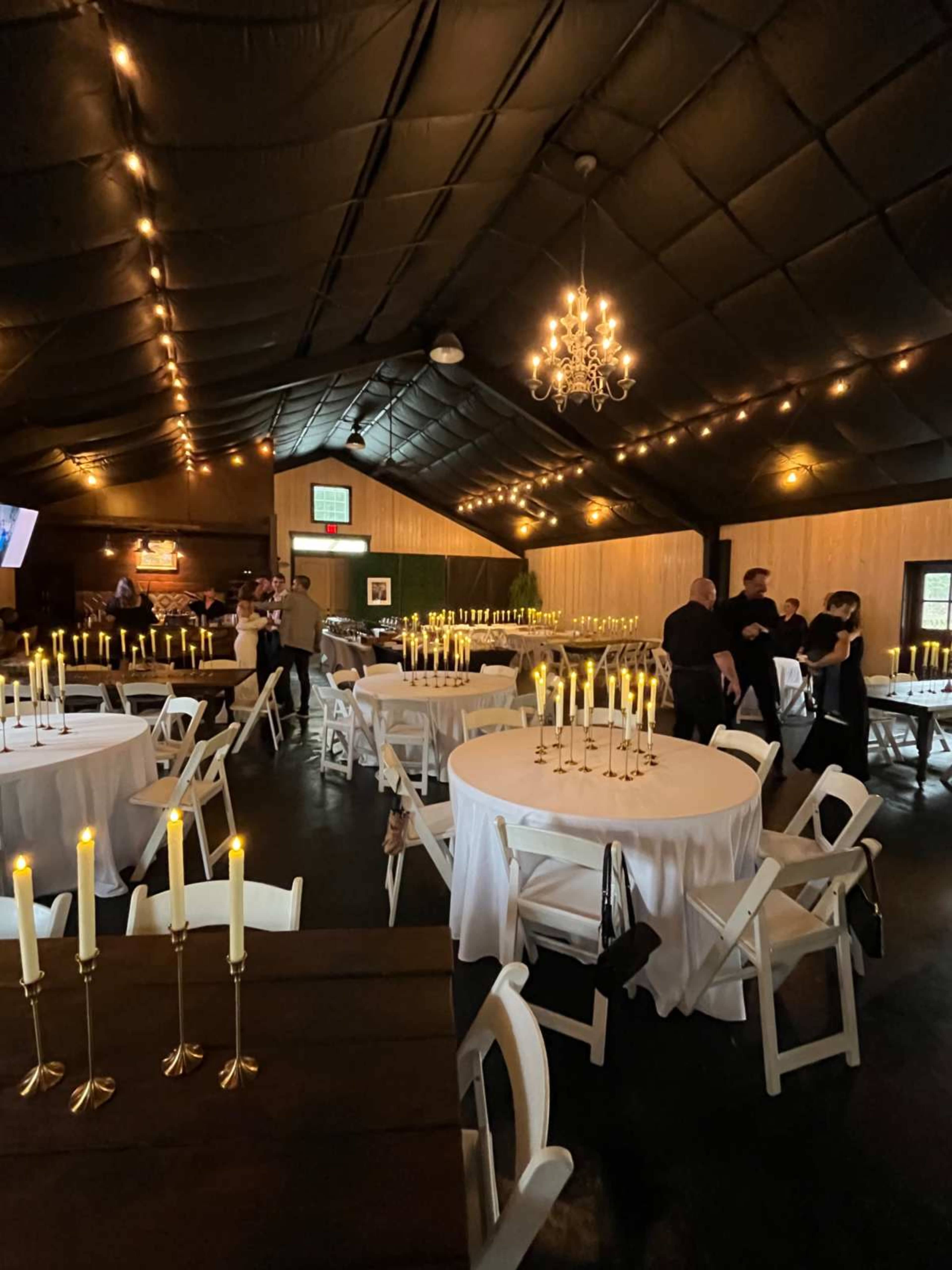 The image shows a dimly lit event space with multiple round tables set for dining, featuring tall candle holders and white tablecloths, while guests socialize in the background.