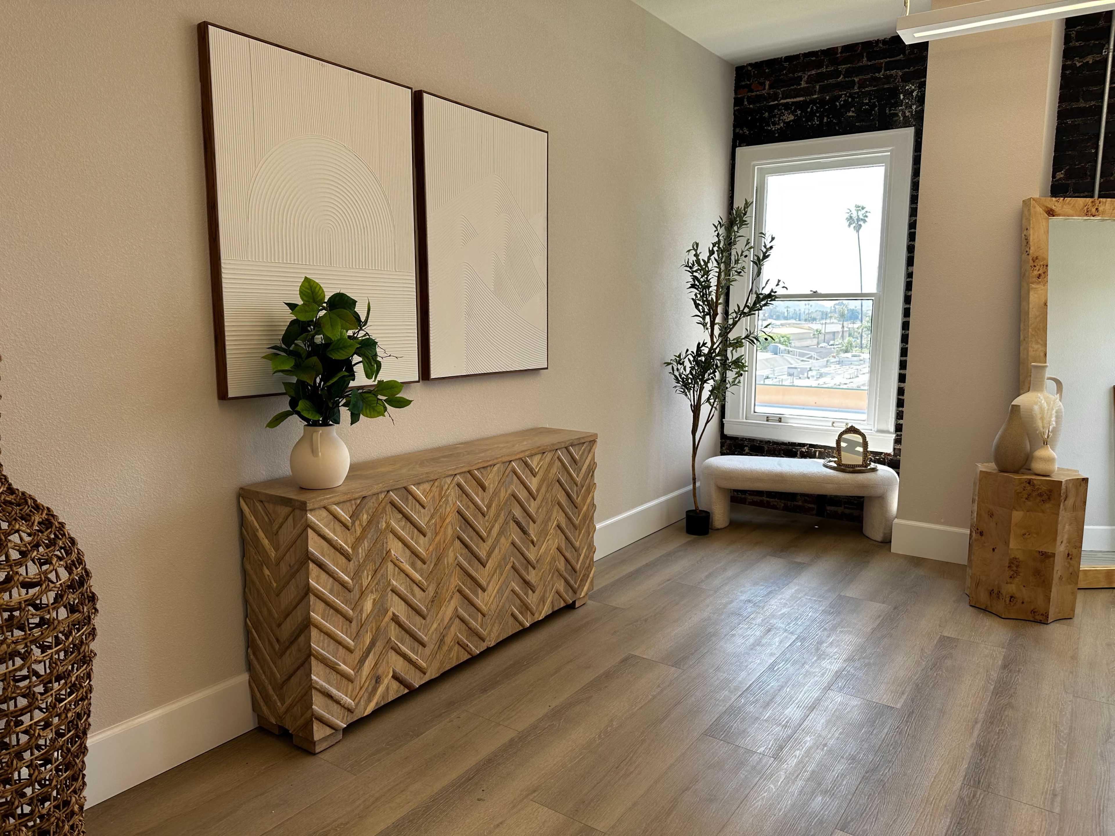 A room with a wooden cabinet featuring a herringbone pattern, two framed artworks on the wall, a potted plant, and large windows with a view of palm trees outside.
