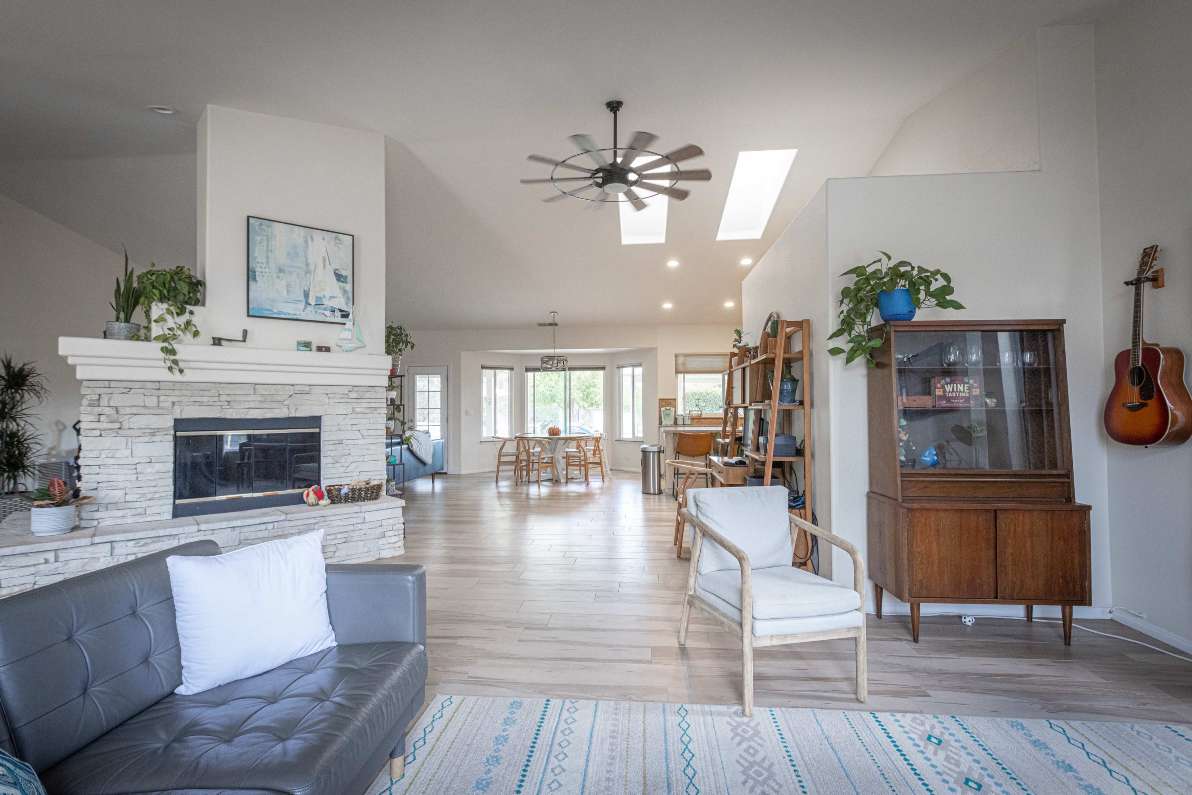 The image shows a spacious living area with a stone fireplace, a gray sofa, and wooden furniture, leading to a bright dining space with large windows.