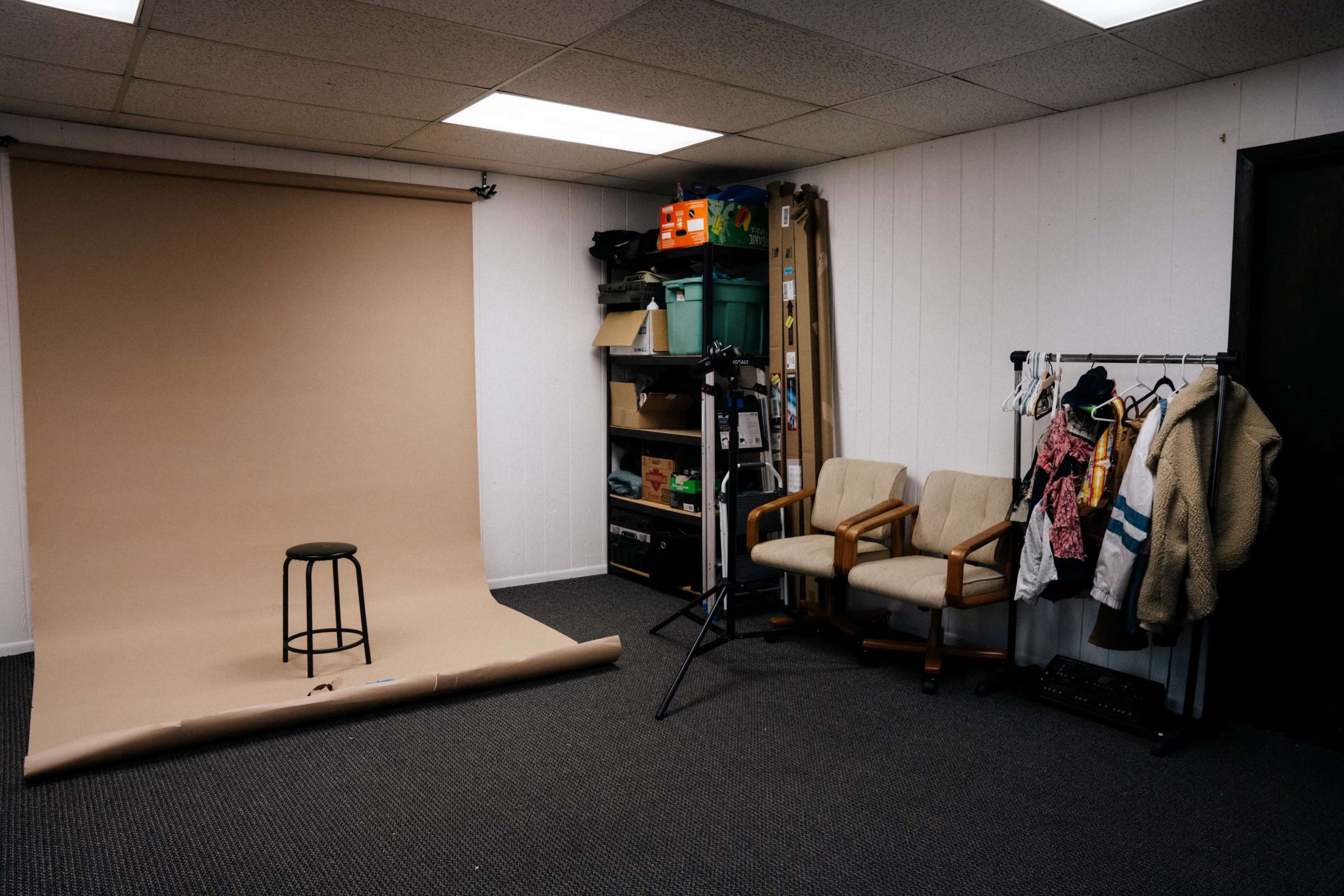 The image shows a photography studio with a beige backdrop, a black stool in the center, two chairs against the wall, and storage shelves filled with various items.
