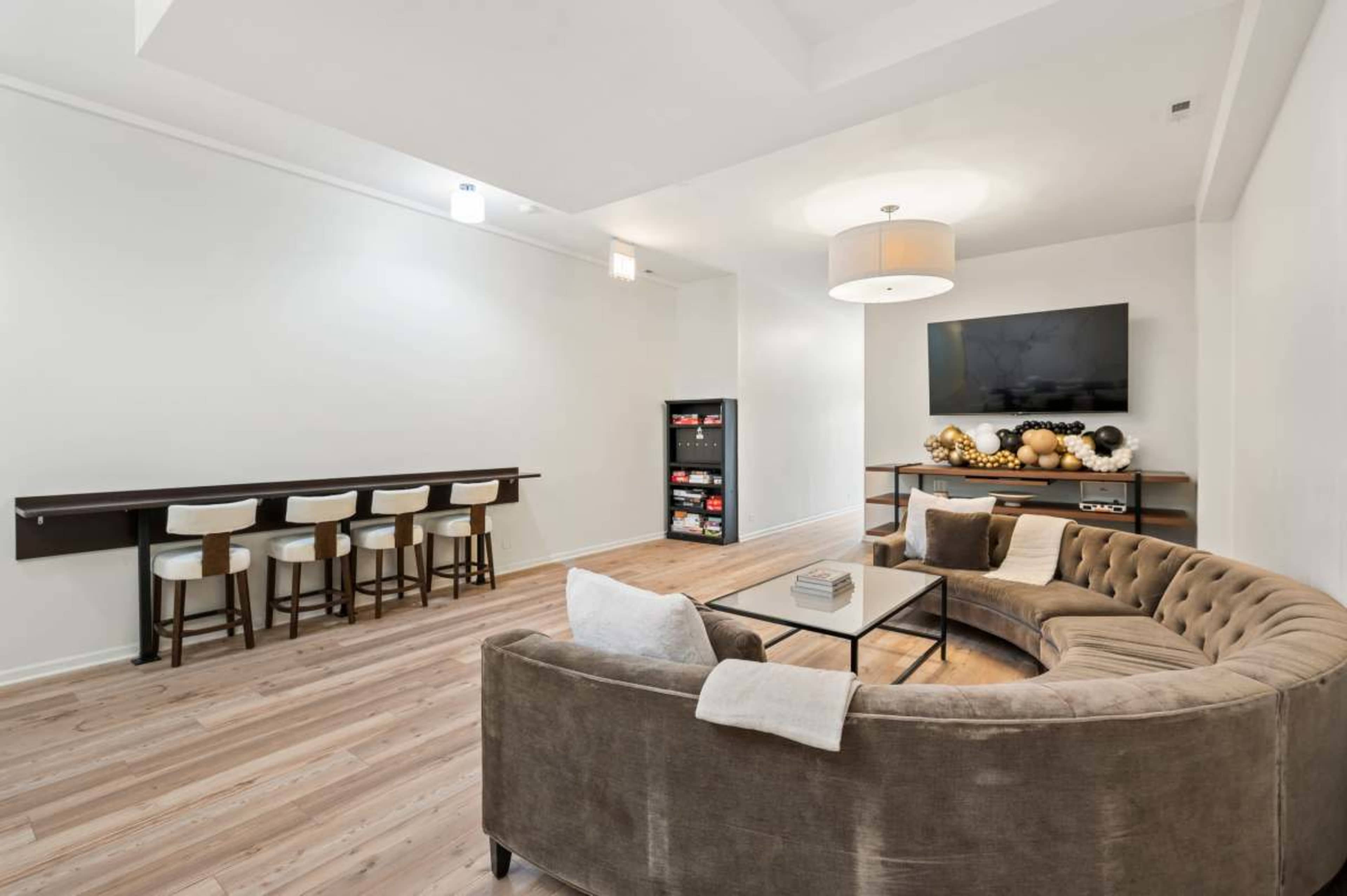 The image shows a modern, minimalist living room featuring a curved sofa, a coffee table, a television mounted on the wall, and a small dining area with bar stools.