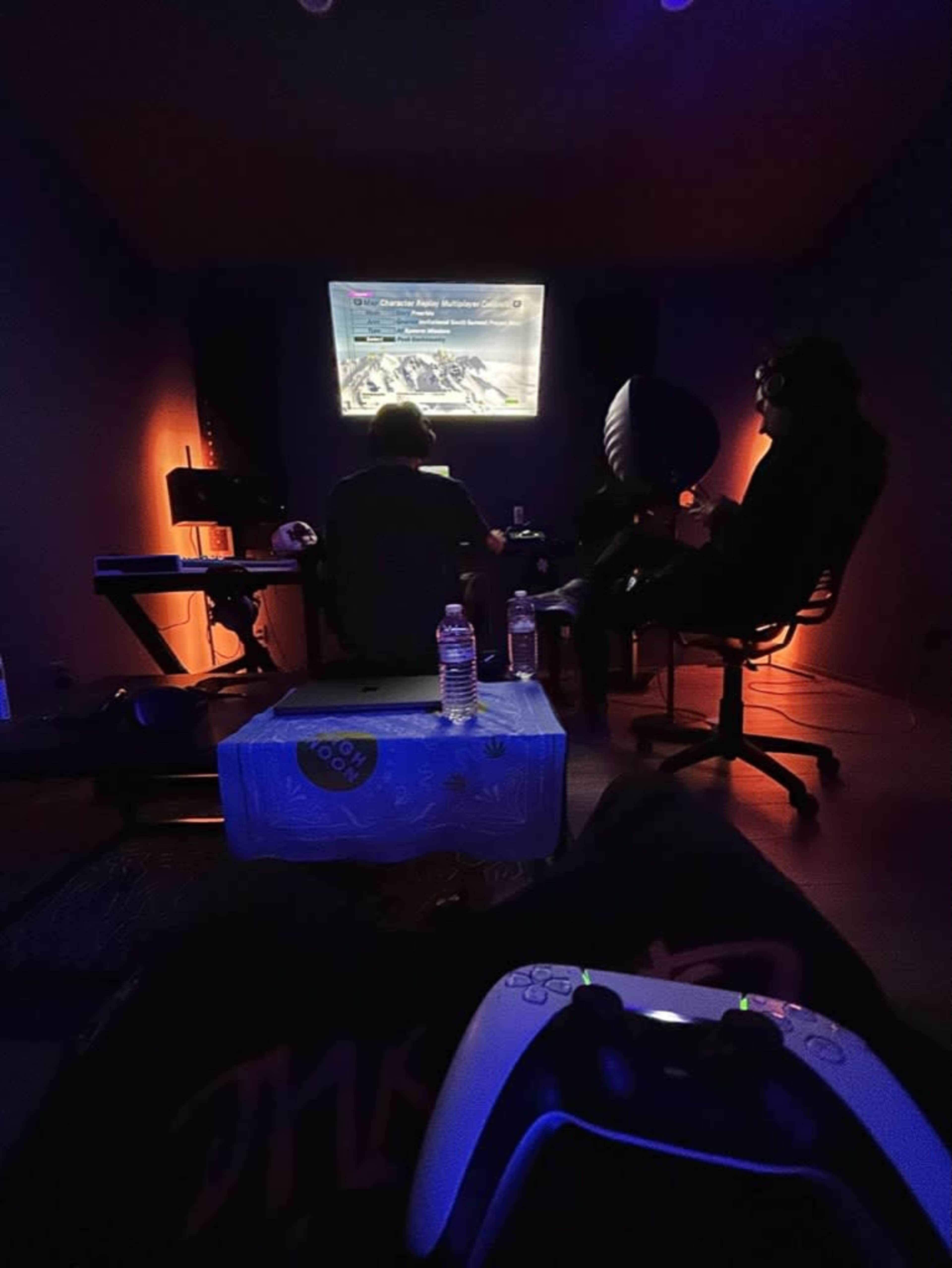 Two people sit in a dimly lit room, focused on a video game displayed on a screen in front of them, with gaming equipment and water bottles nearby.