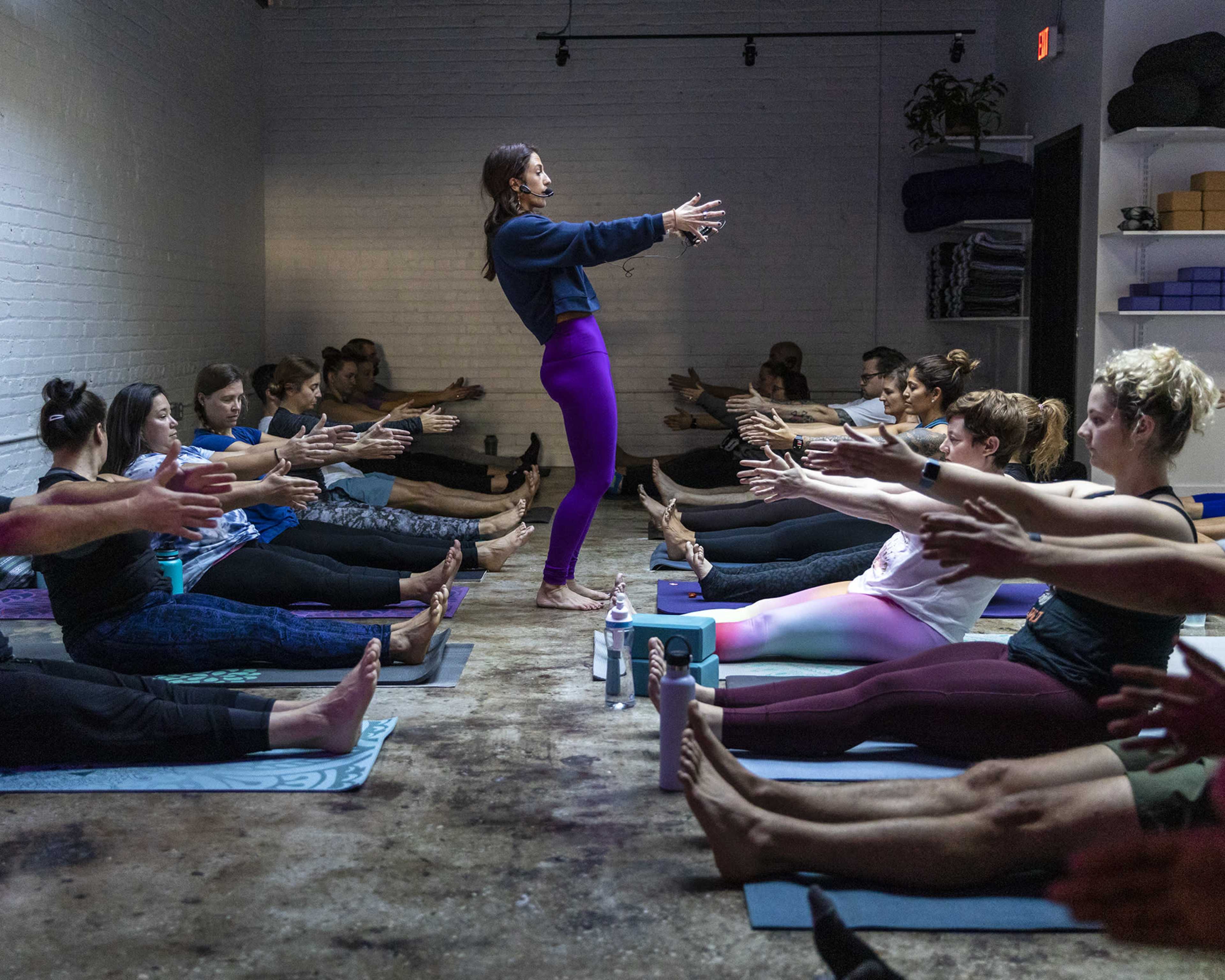 A yoga instructor demonstrates a pose in front of a class of students seated on mats, all reaching forward with their arms extended.