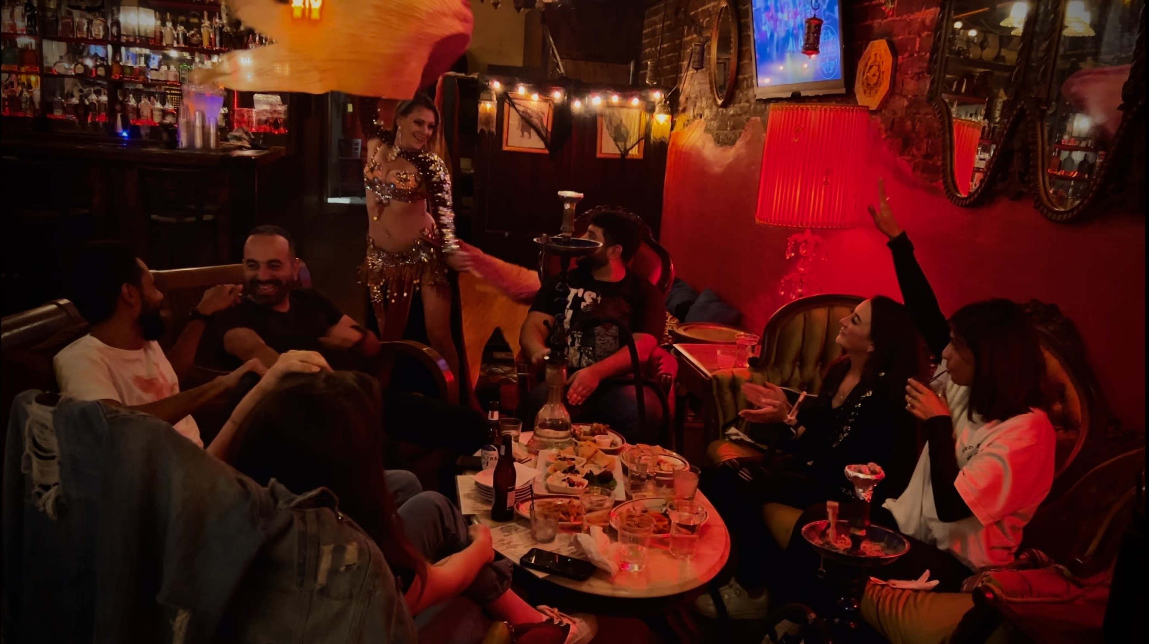 A group of people gathers around a table filled with food and drinks while a performer in a sequined costume entertains them in a dimly lit room adorned with colorful decorations.