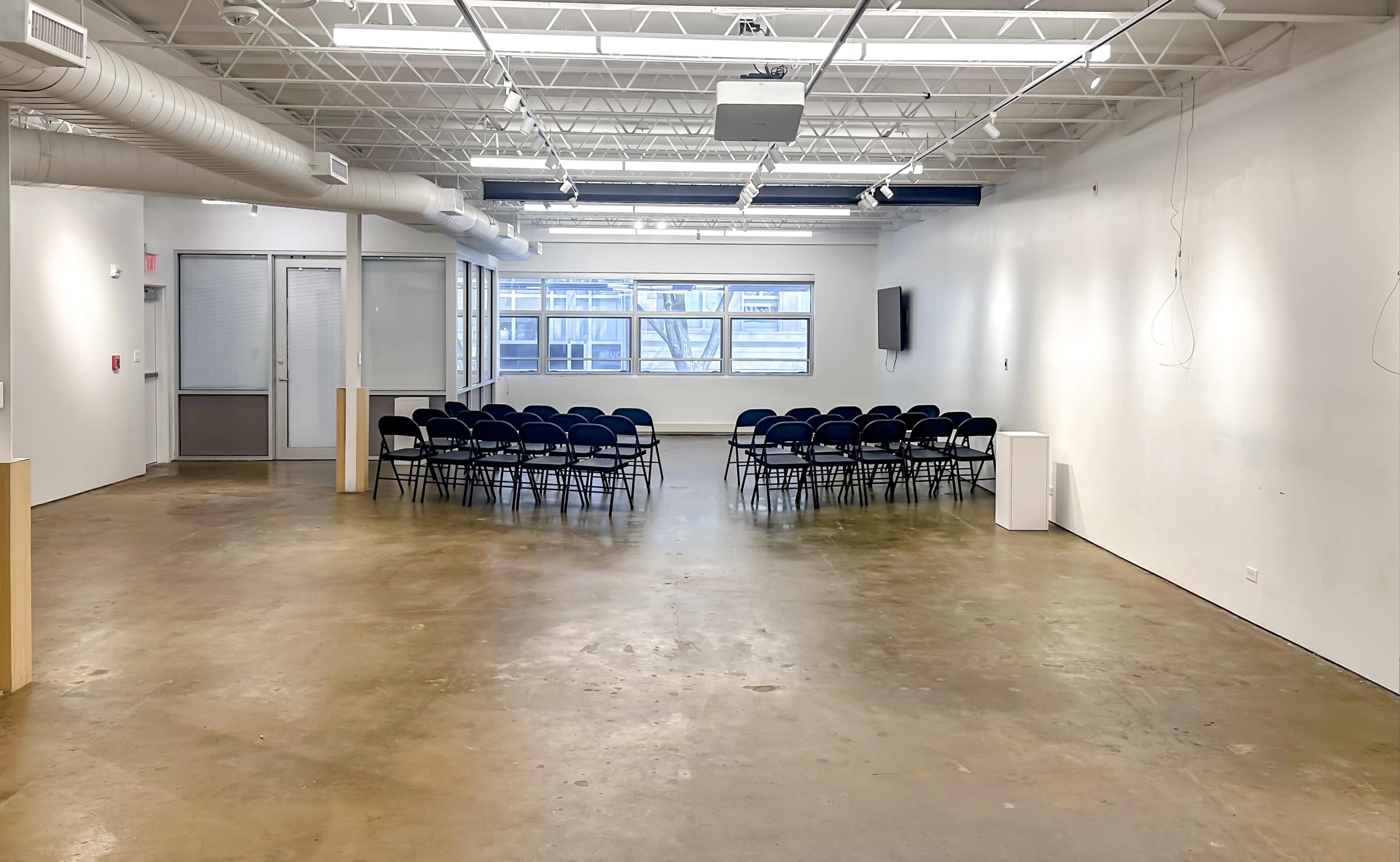 A large, empty room with a concrete floor and rows of black chairs arranged for an event.
