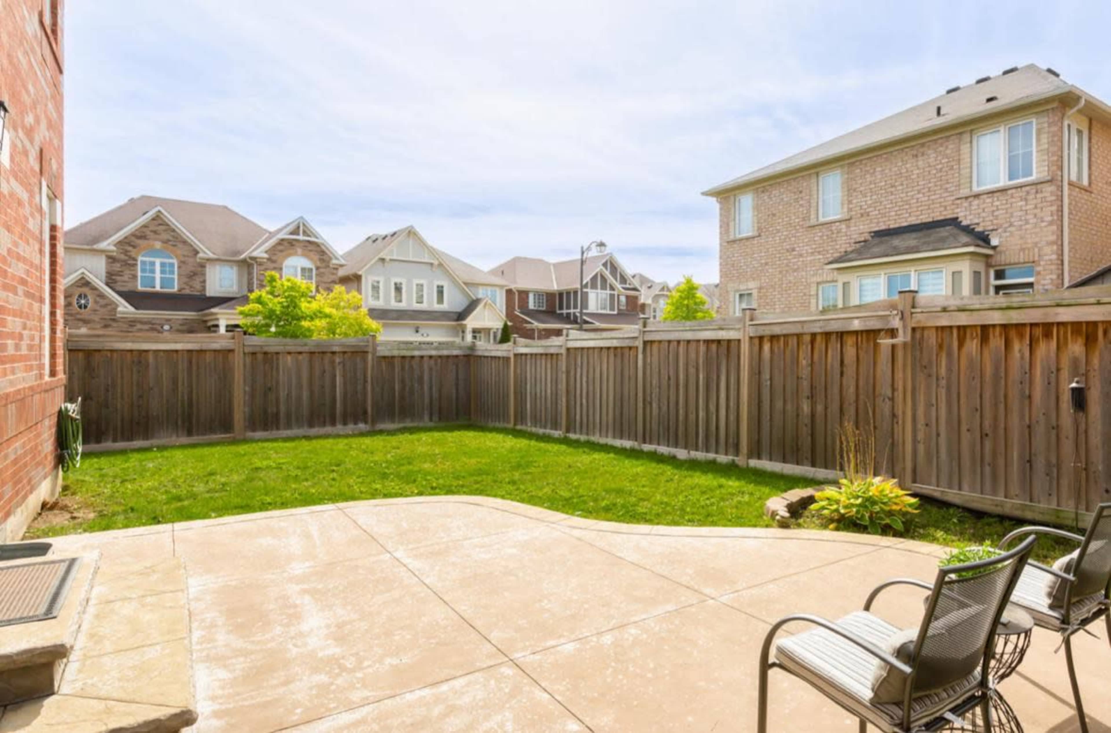 The image shows a backyard with a grassy area surrounded by a wooden fence and two chairs on a concrete patio.