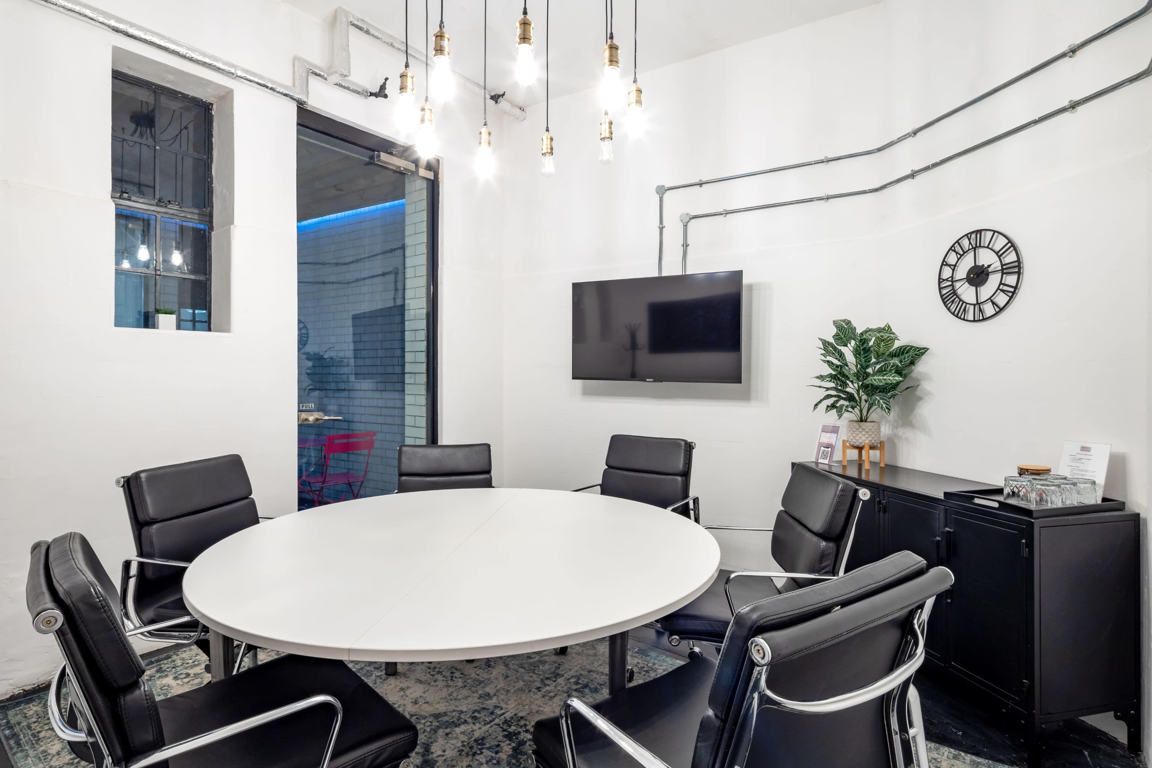 A modern conference room features a round white table surrounded by black leather chairs, a flat-screen TV on the wall, and industrial-style lighting above.