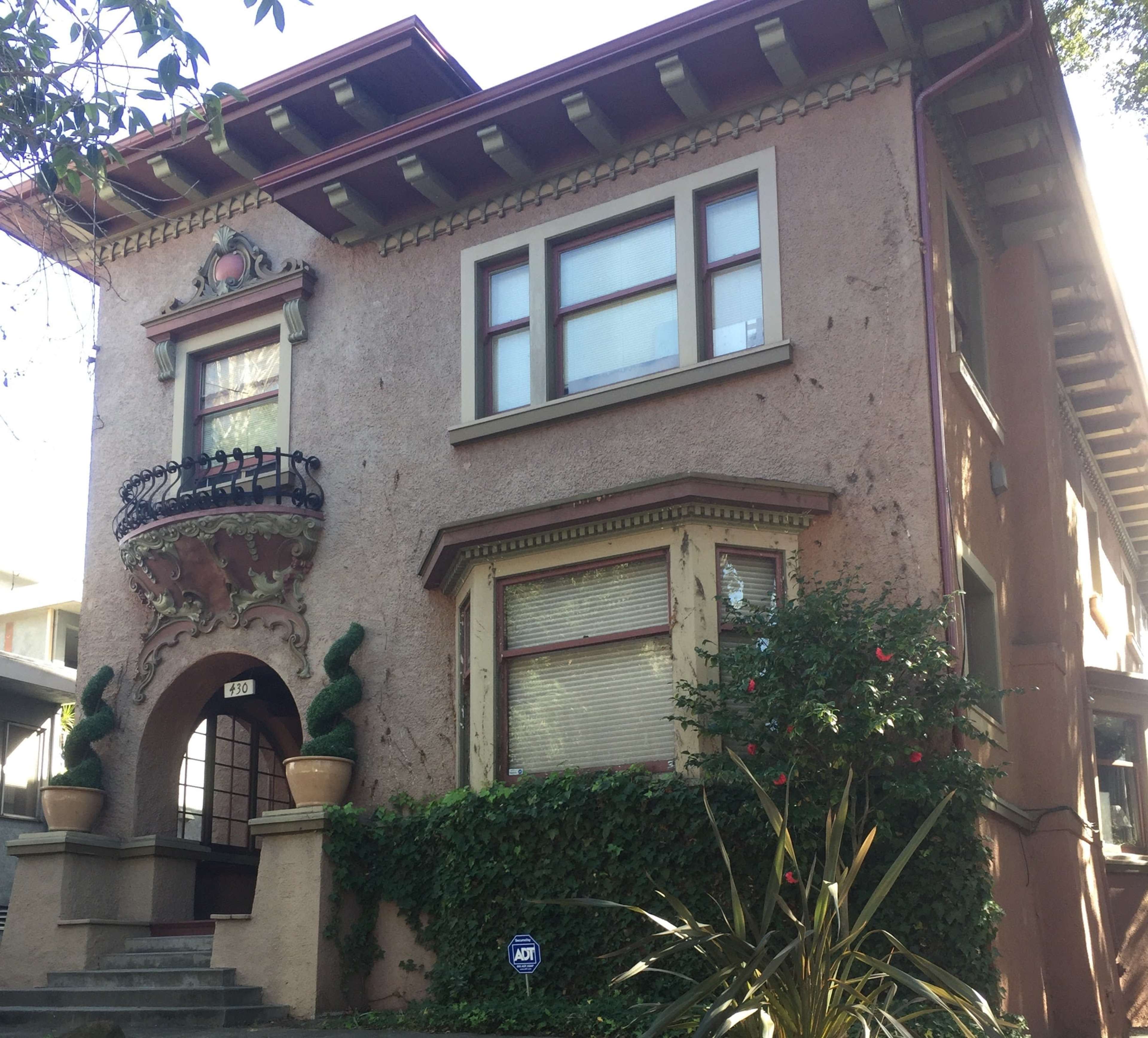 The image shows a two-story, decorative apartment building with a front balcony and climbing plants around its entrance.