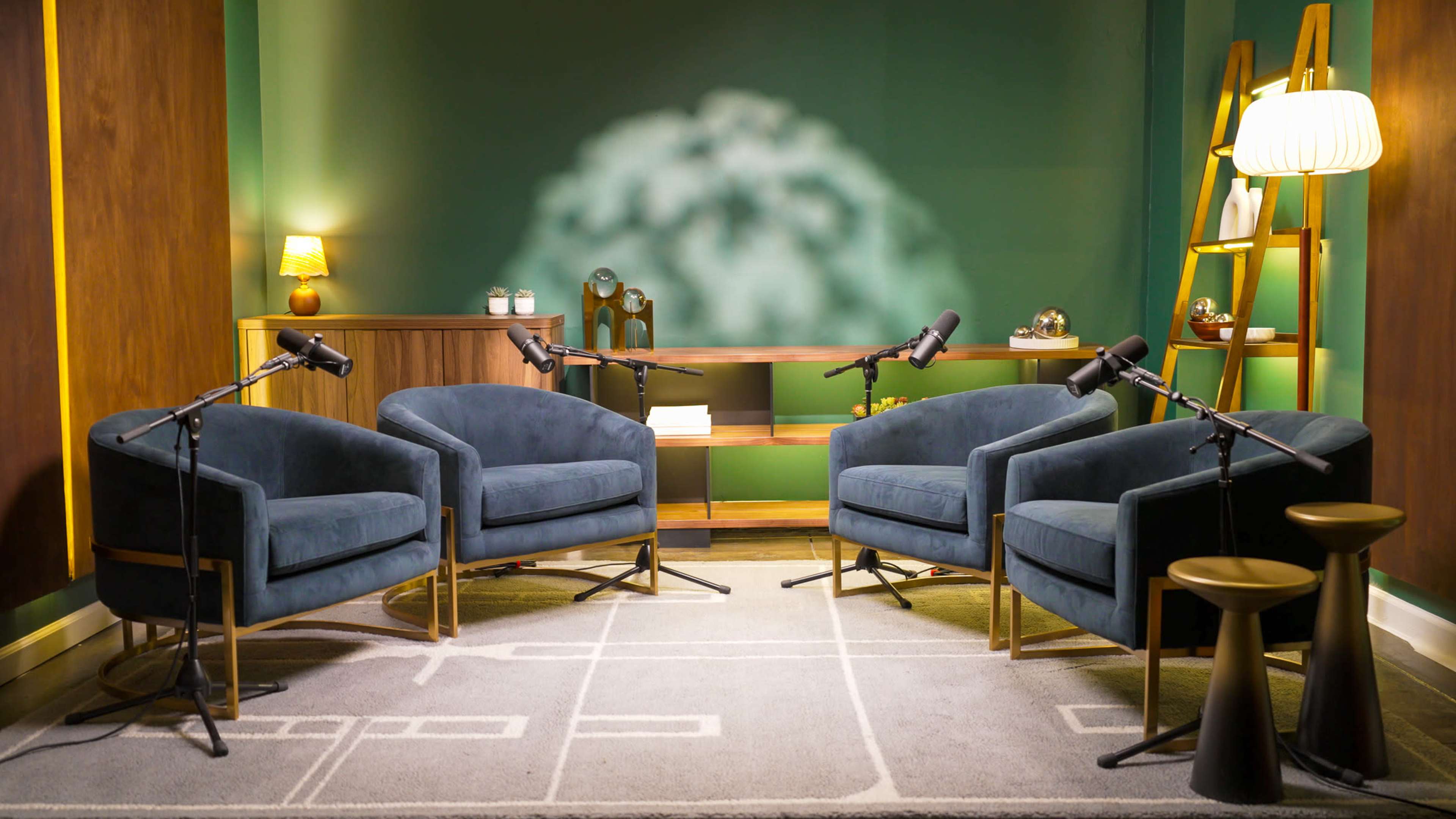 The image shows a cozy interview setup featuring three blue armchairs and a two-tiered wooden shelf, all arranged in a softly lit room with a green backdrop.