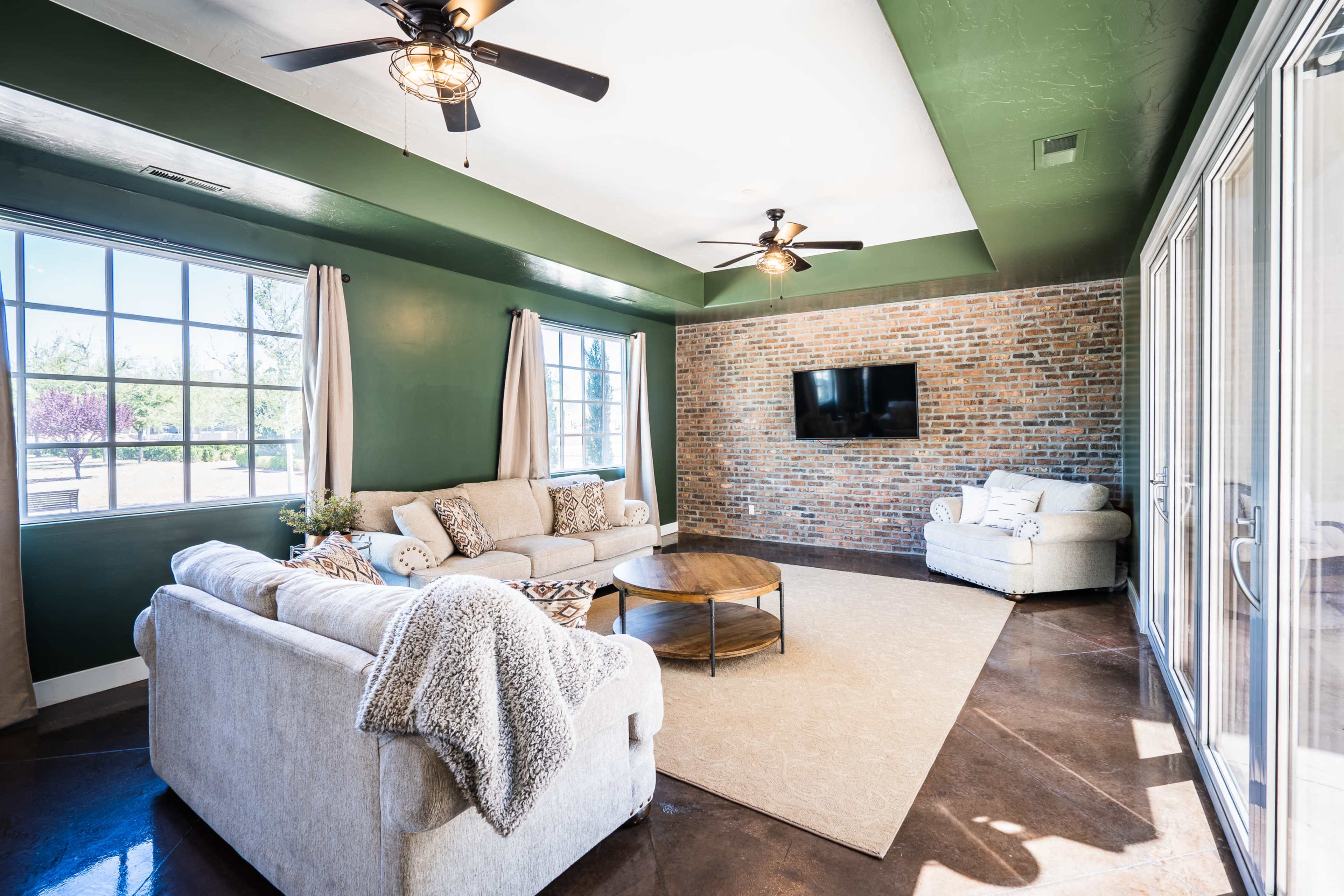 A living room with a green ceiling and exposed brick wall, featuring a beige sofa set, a round coffee table, and large windows.