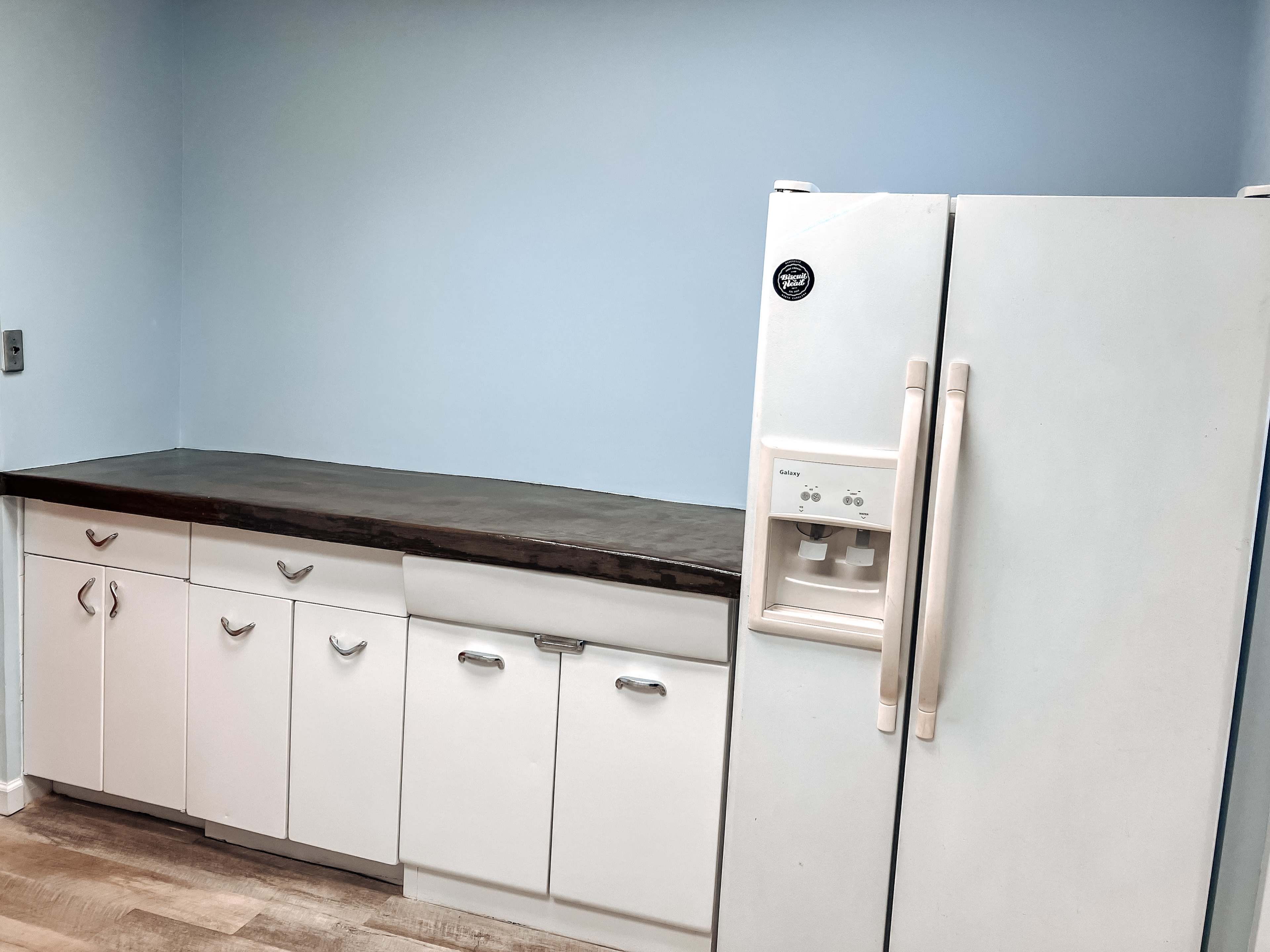 A kitchen space features a blue wall, a countertop, and white cabinetry with a side-by-side refrigerator.
