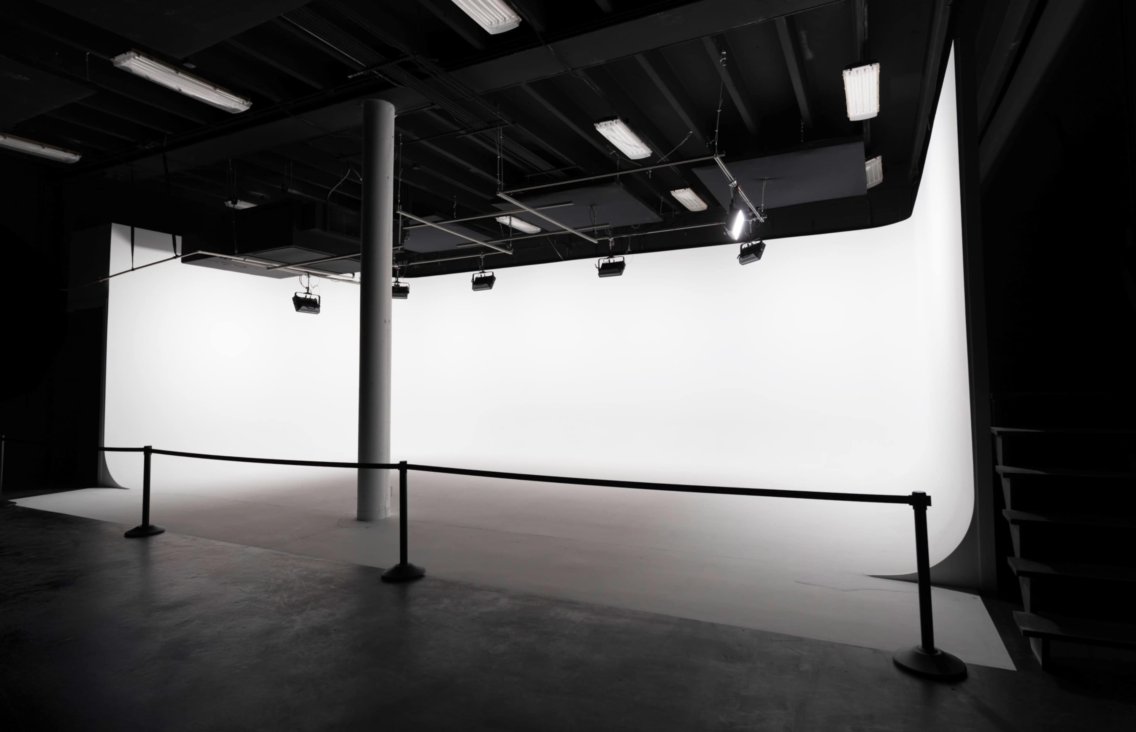 The image shows a photography studio with a seamless white backdrop, illuminated by overhead lights, and enclosed by a rope barrier.