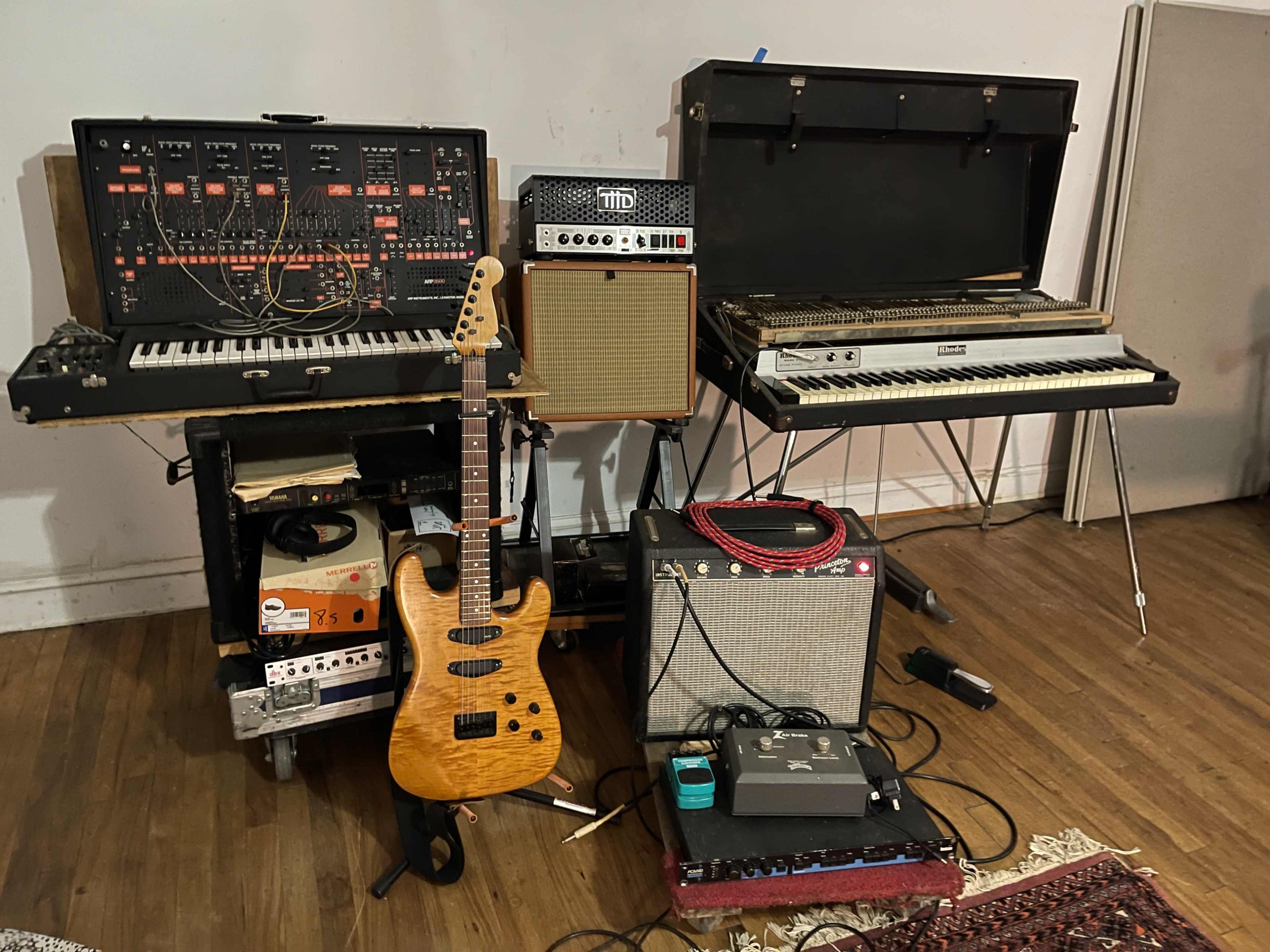 A setup of various musical equipment, including synthesizers, amplifiers, a guitar, and effects pedals, arranged on a wooden floor.