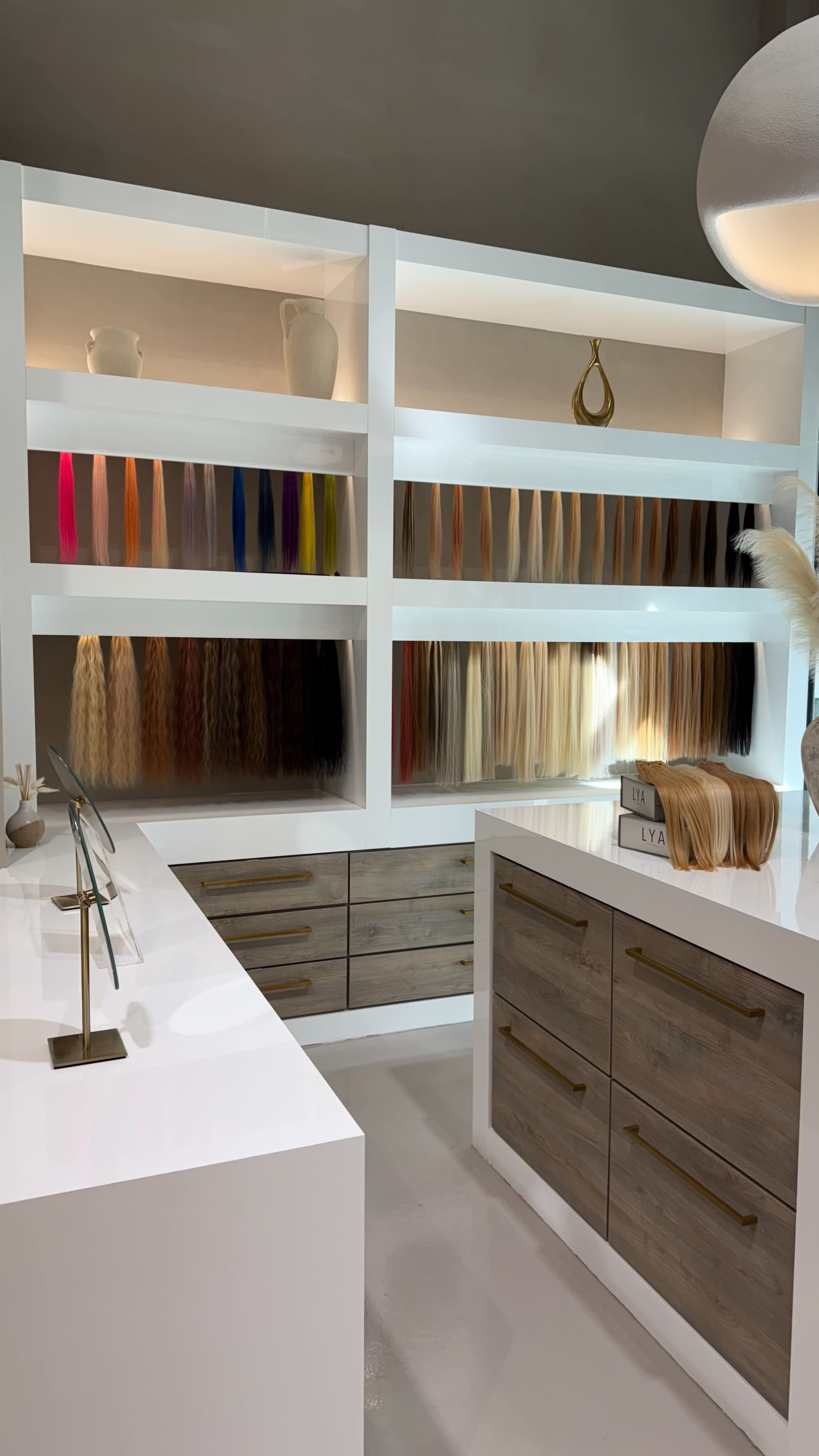 The image shows a modern hair salon interior featuring neatly organized hair extensions in various colors displayed on shelves, with a sleek countertop and decorative elements.