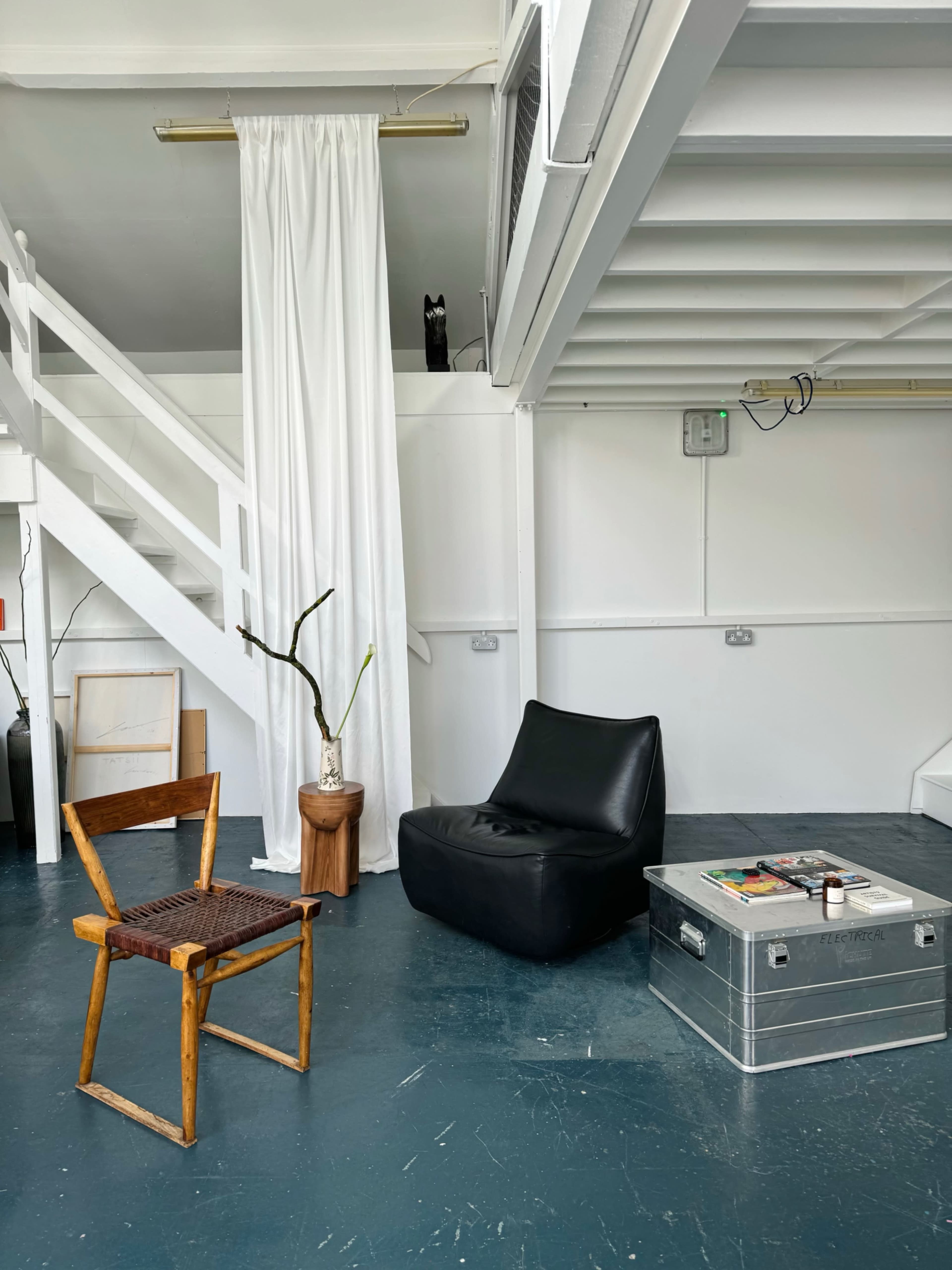 The image shows a minimalist interior featuring a wooden chair, a black lounge chair, a metal box serving as a coffee table, and a vase with a plant, all within a space that includes a staircase and white curtains.