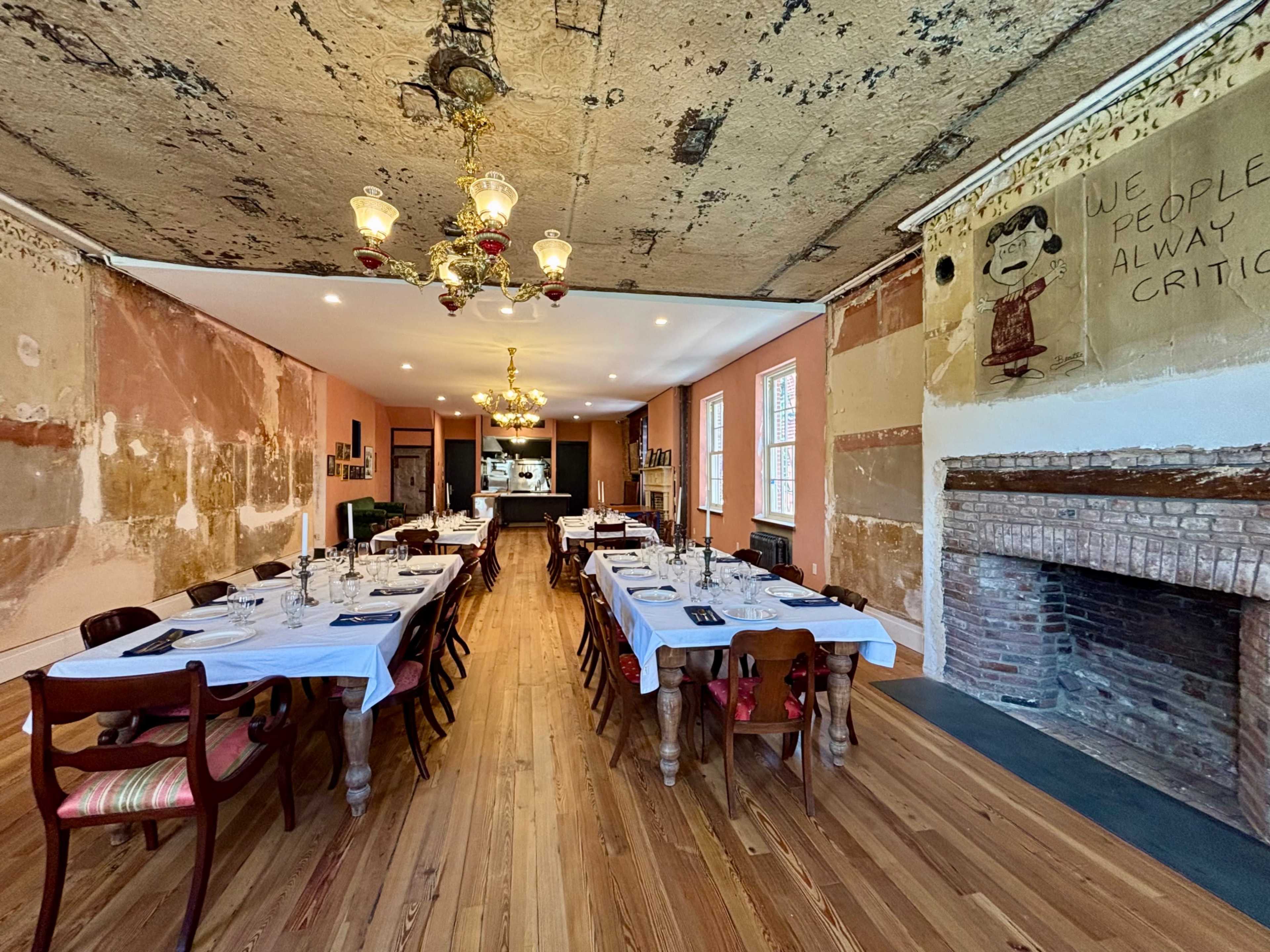 The image shows a spacious dining room with a long, set dining table, wooden floors, exposed brick fireplace, and walls partially covered with peeling paint and graffiti.