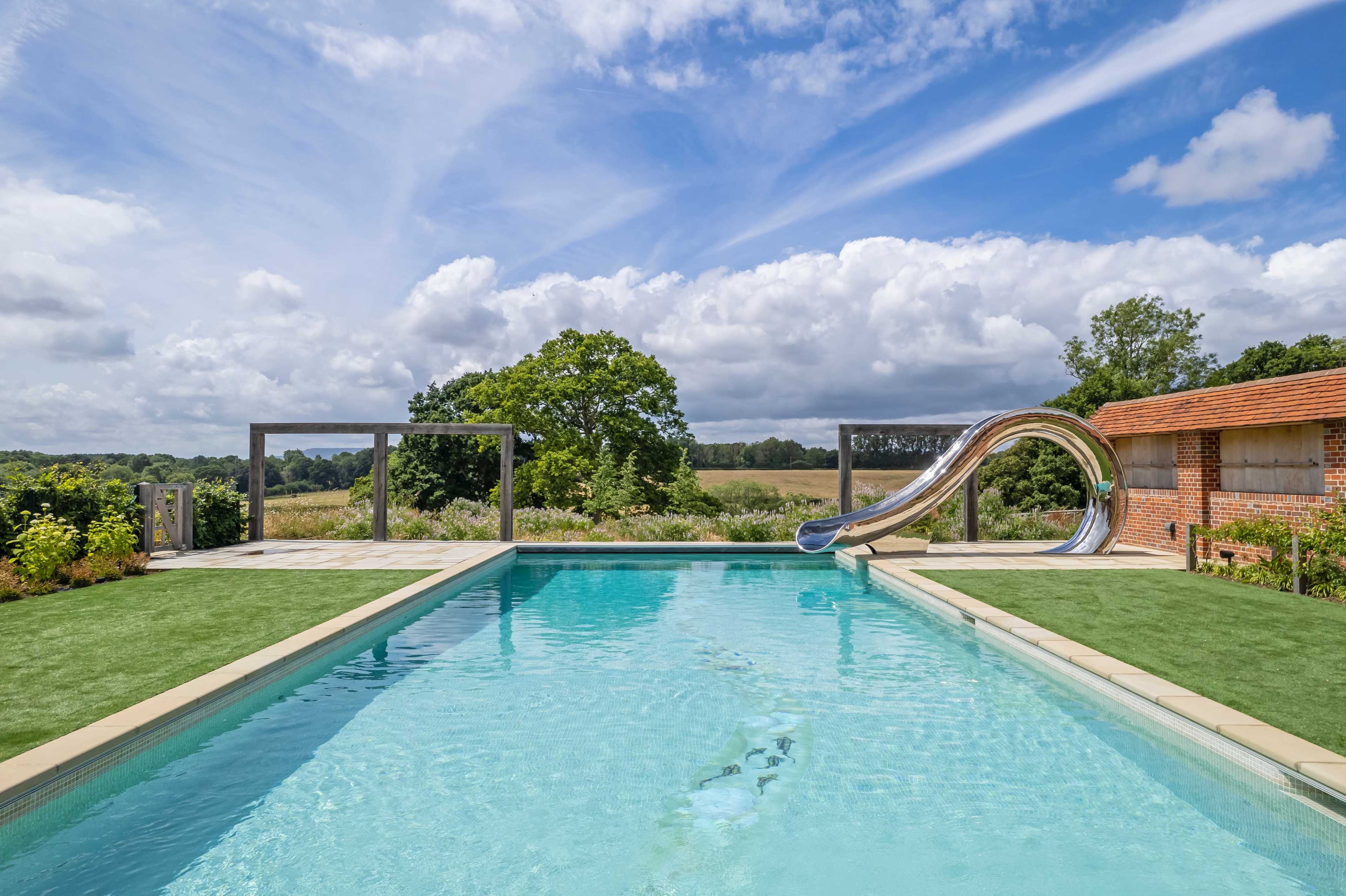 A modern swimming pool with a sliding water feature is surrounded by green grass and overlooks a rural landscape under a partly cloudy sky.