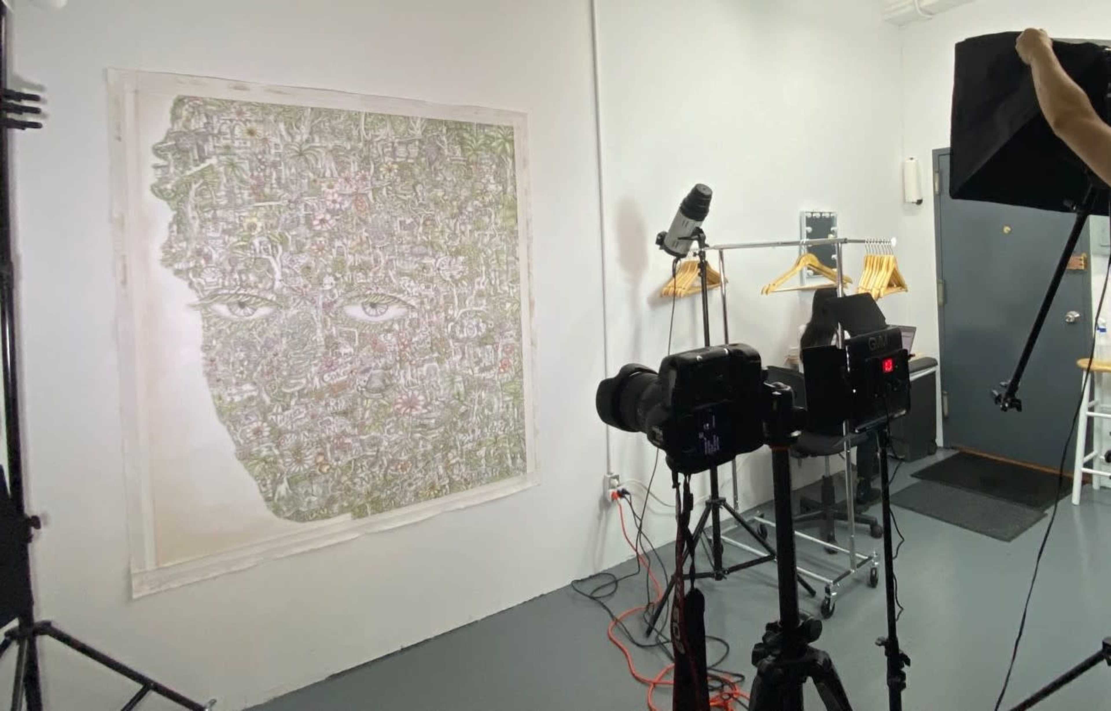 The image shows a gallery space with a large artwork depicting a face created from various symbols, surrounded by photography equipment and a clothing rack.