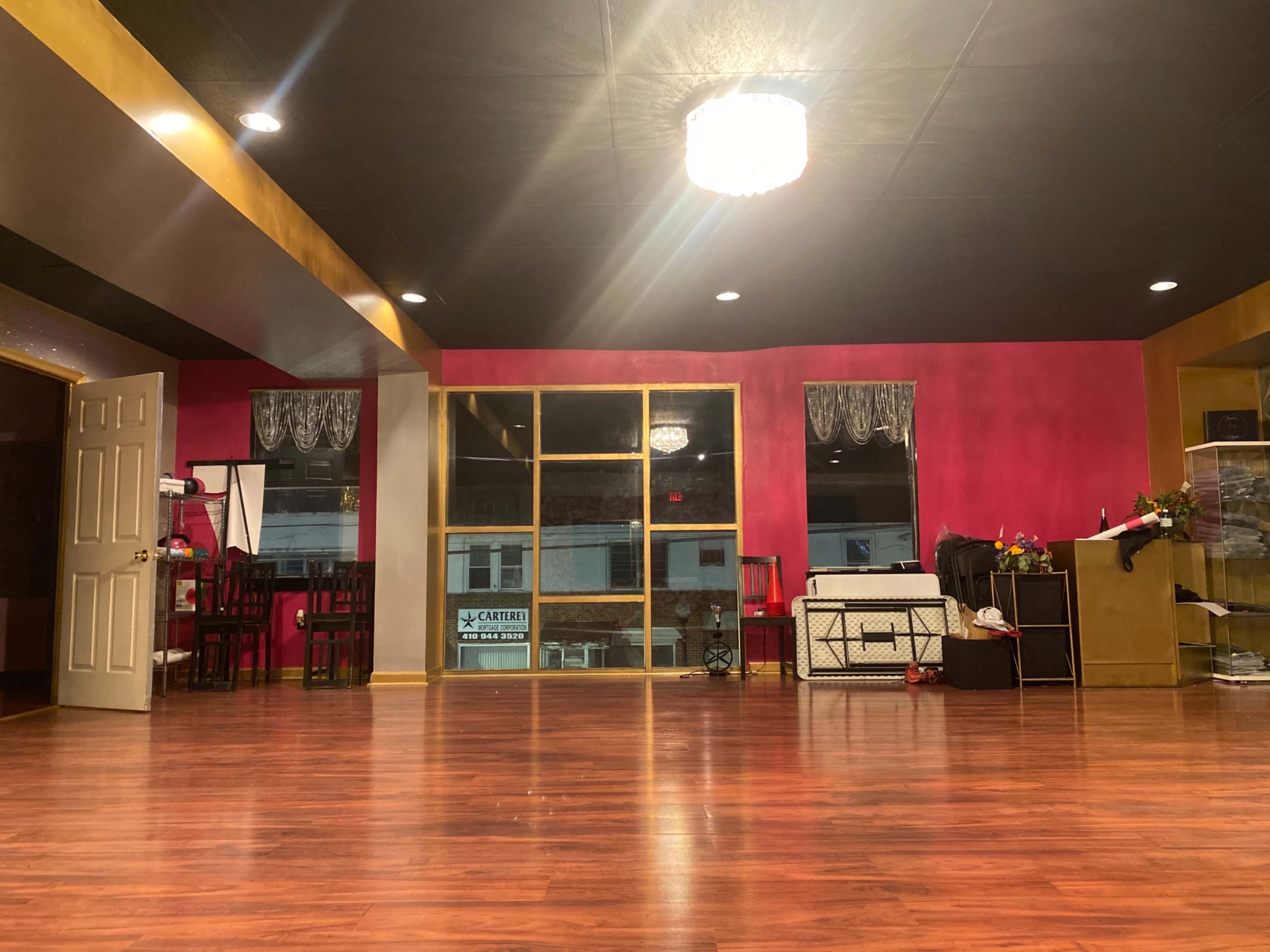 A spacious dance studio with a polished wooden floor and pink walls, featuring large windows and various pieces of furniture along the edges.