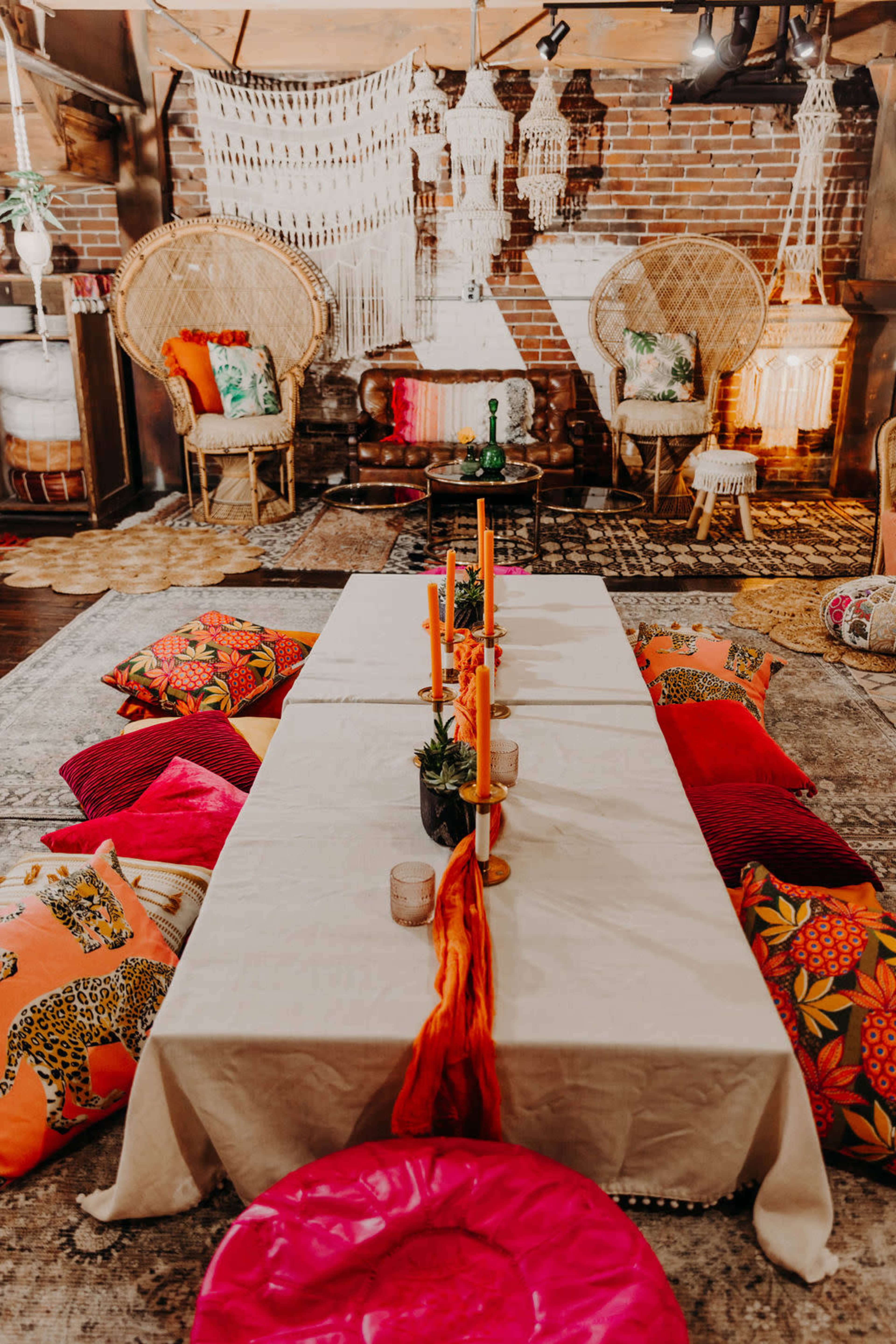 A decorated indoor space featuring a long table set with candles and surrounded by vibrant cushions and two large, woven chairs.