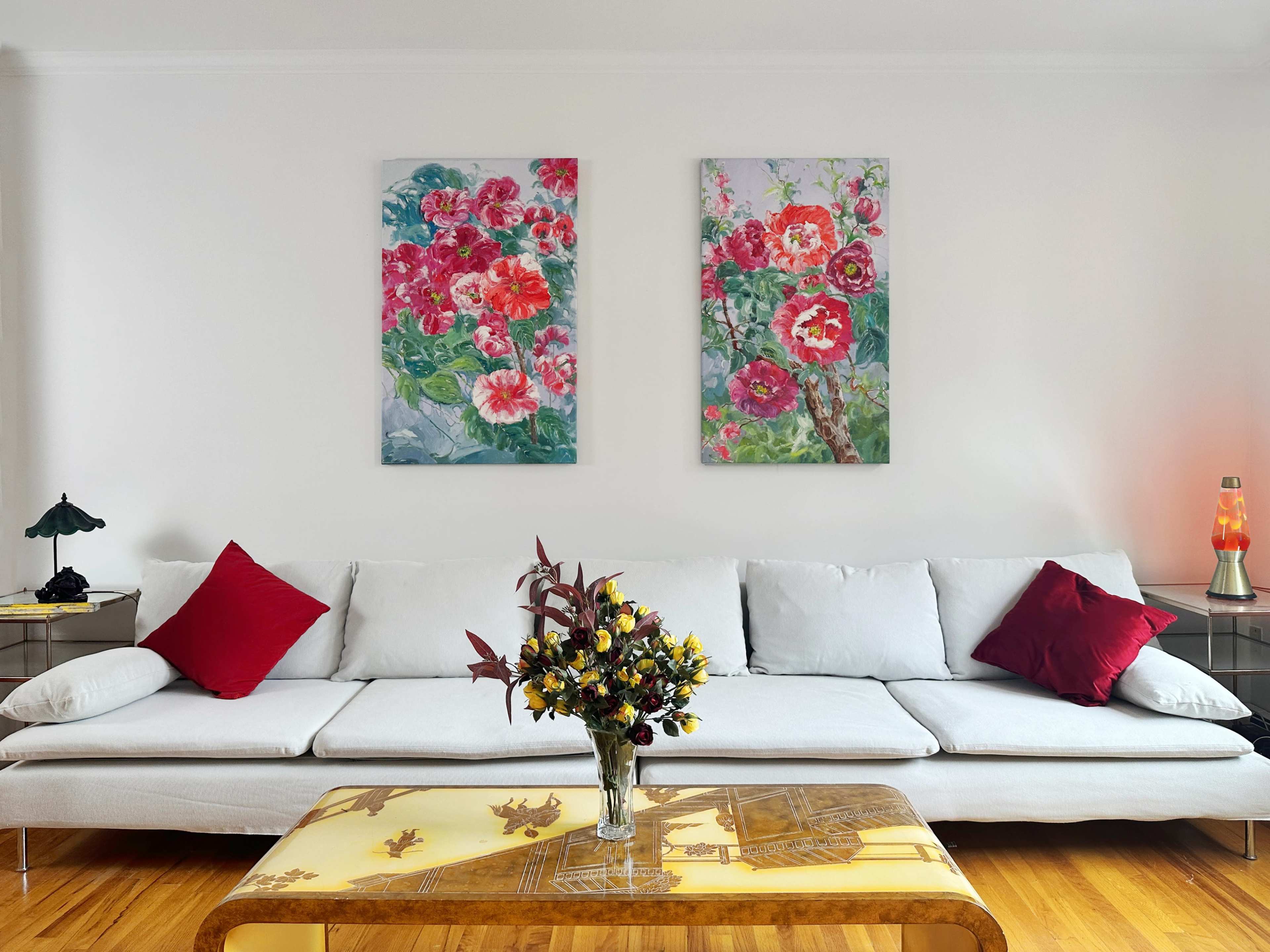 A modern living room features a white sofa with red cushions, a glass coffee table, and two floral paintings on the wall.