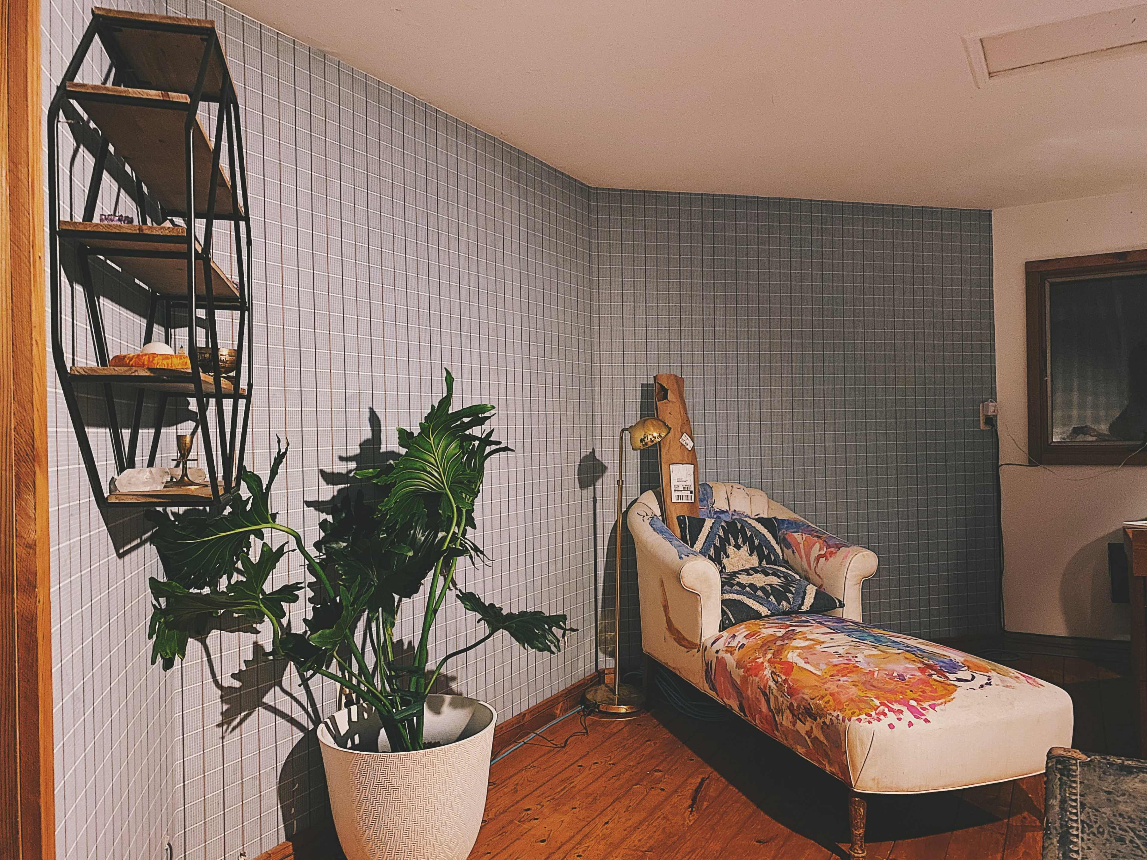 The image shows a cozy room with a patterned chaise lounge, a potted plant, and a shelf mounted on the wall displaying decorative items.