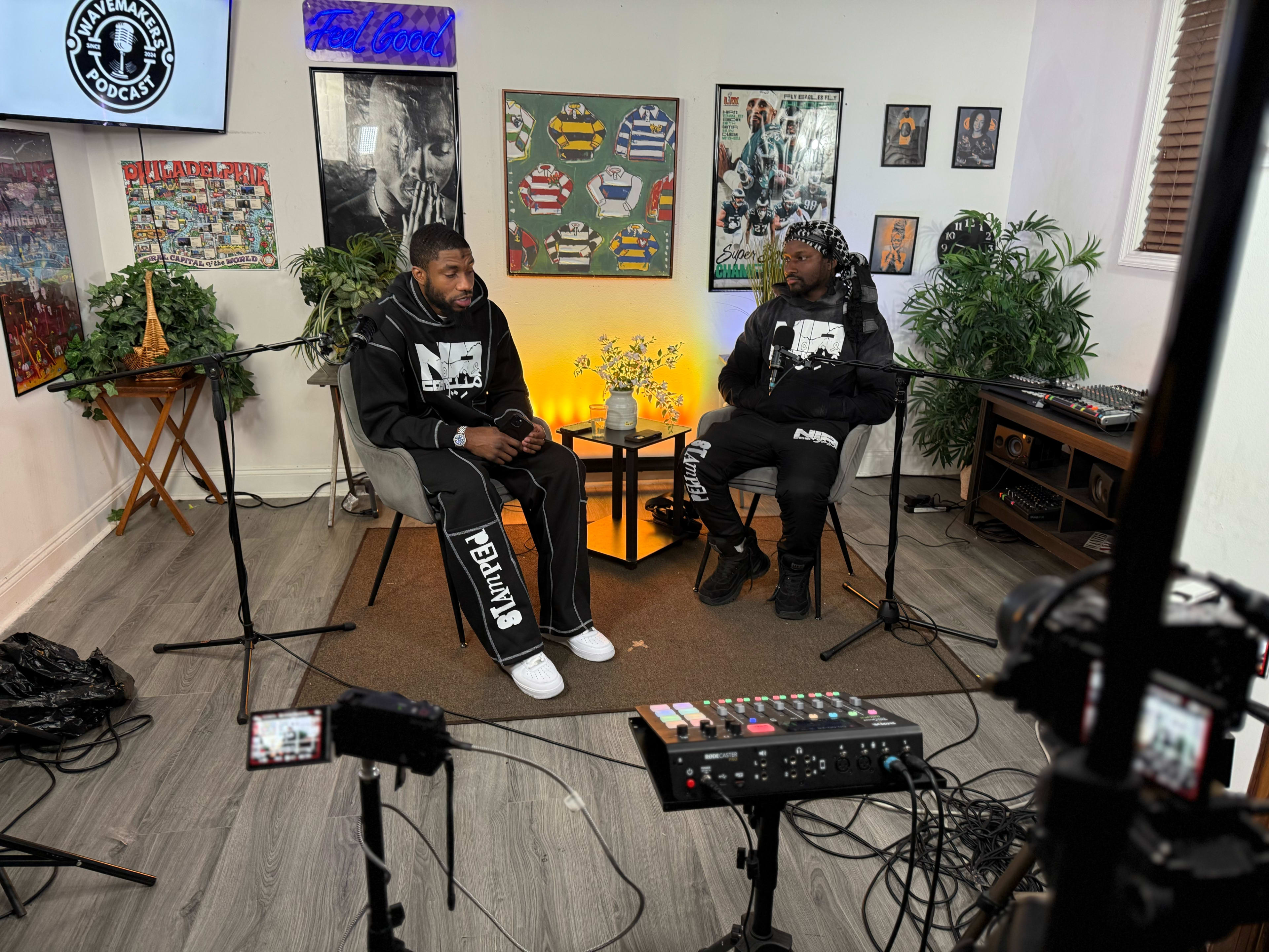 Two men sit in a podcast studio surrounded by various wall art and plants, engaged in conversation while cameras and microphones are positioned around them.